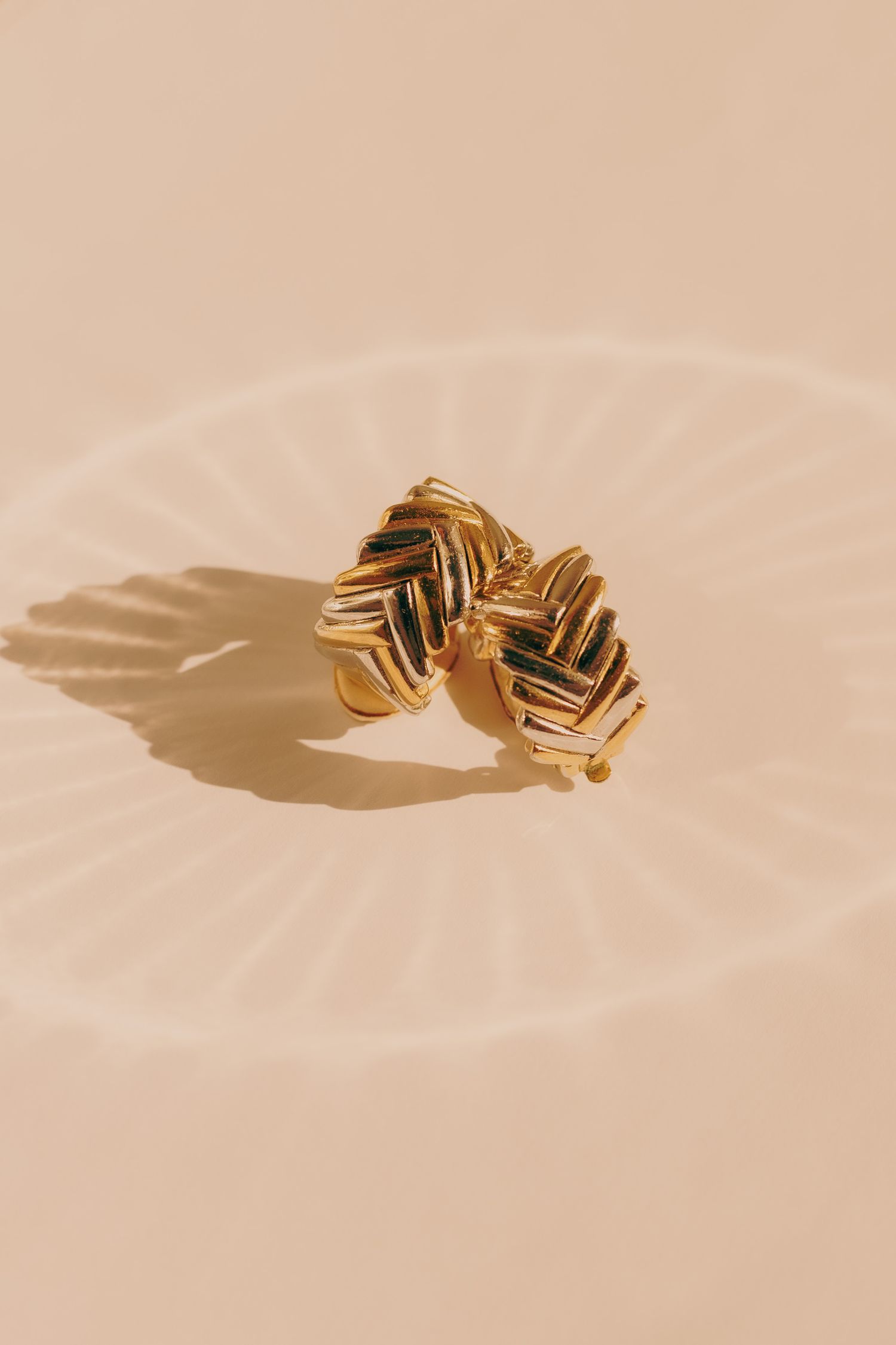 Two gold woven chevron earrings leaning together on a beige surface with circular shadow.