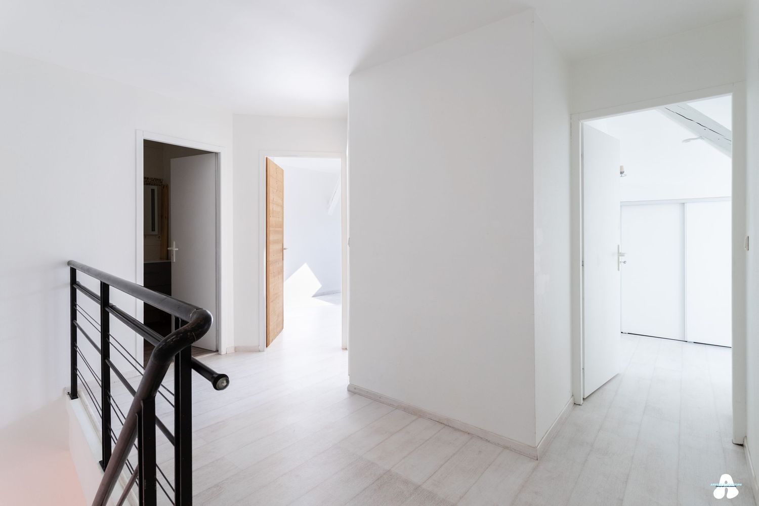 Bright white hallway with black metal stair railing and light wood flooring leading to multiple rooms.