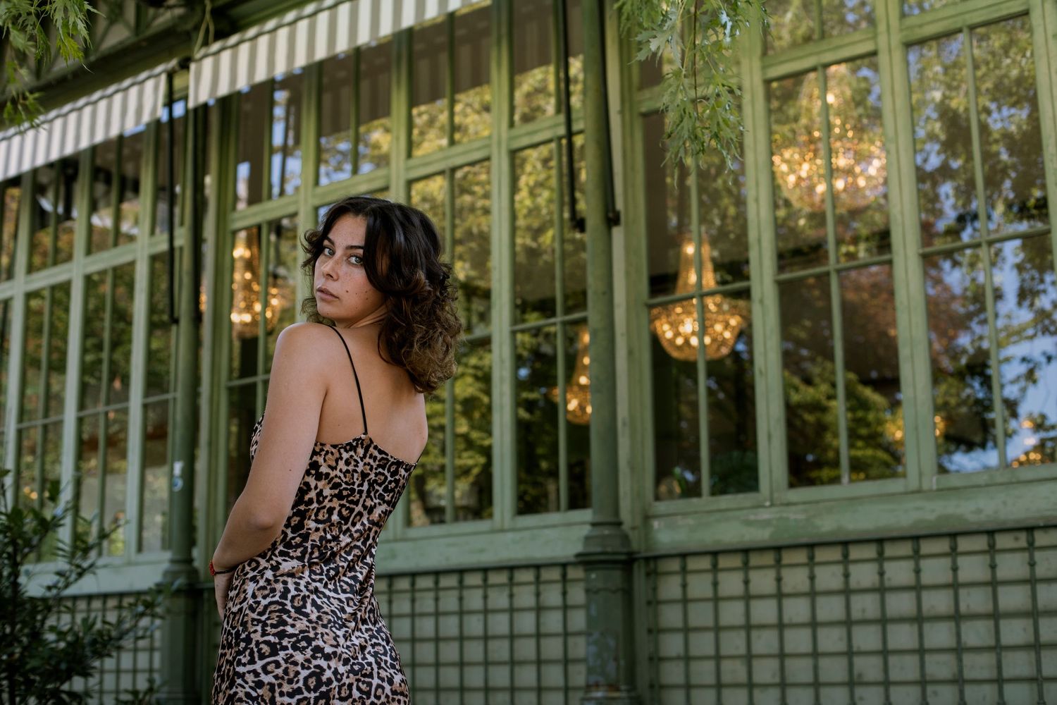 Woman in leopard print dress looking back over shoulder near a green greenhouse window.