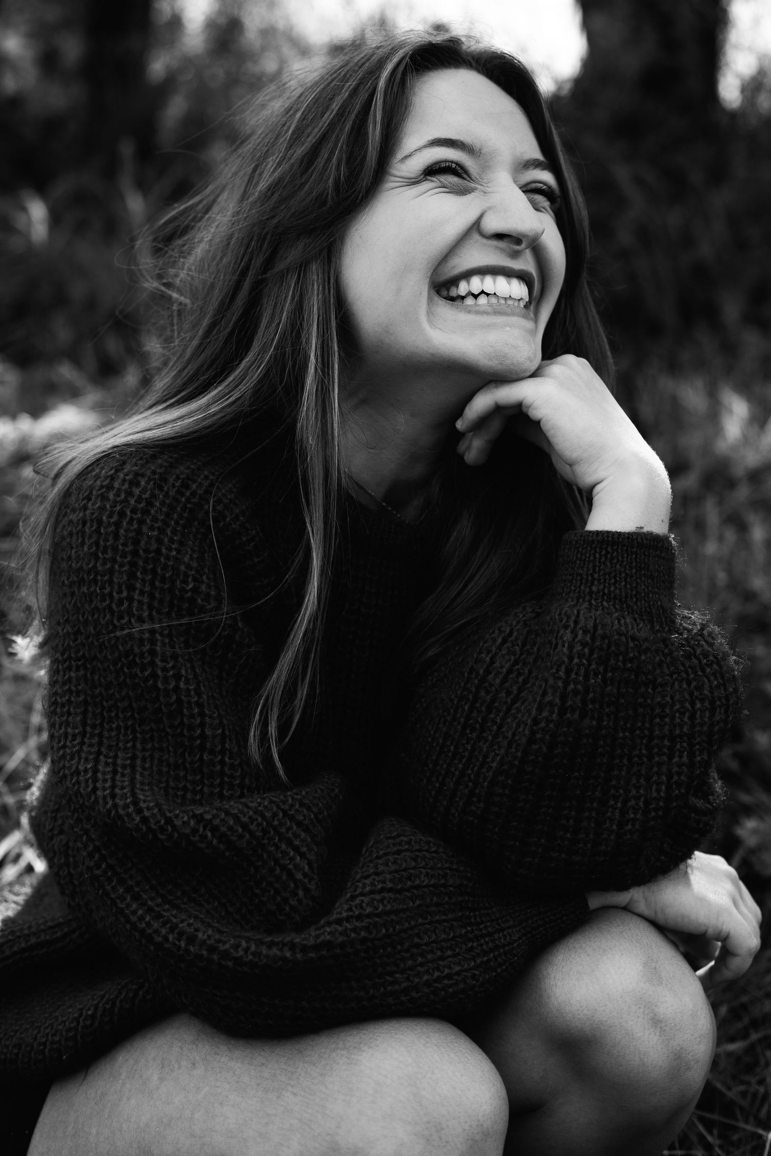 Black and white portrait of a laughing woman in knit sweater sitting outdoors among trees.