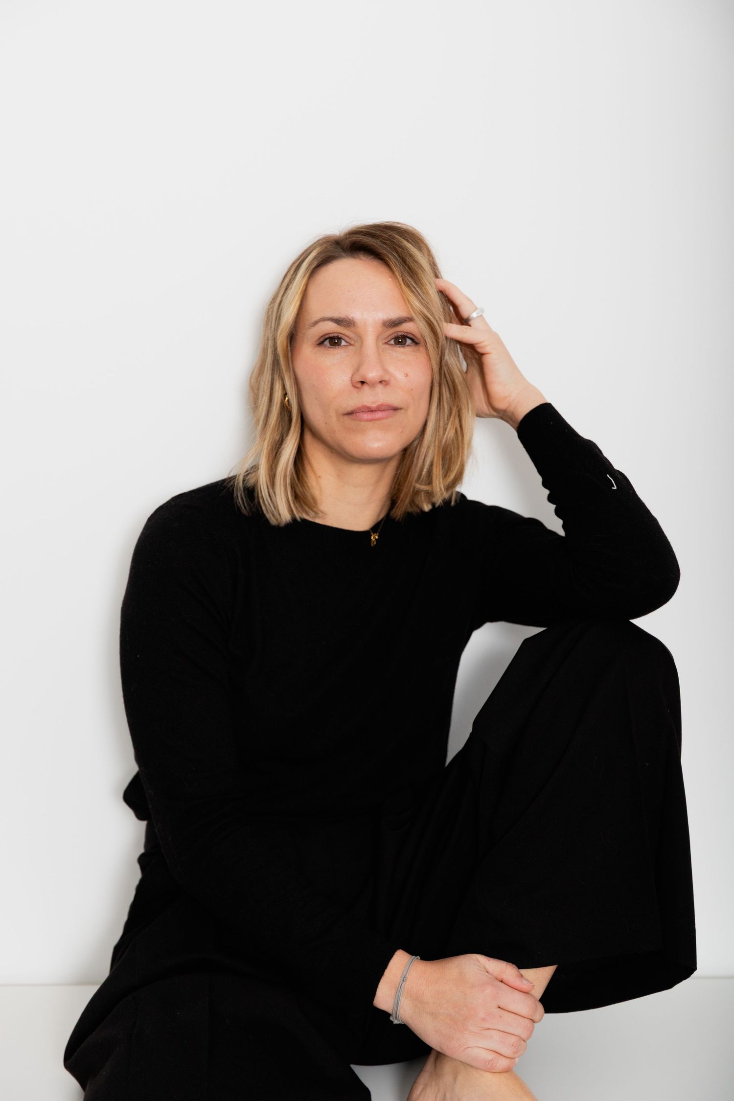 Blonde woman in black outfit sitting on floor against white wall, casual portrait photo.