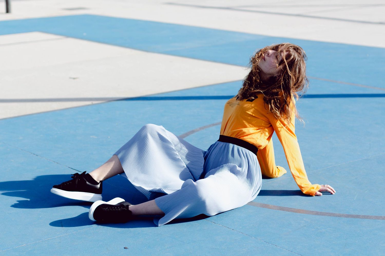 Woman in yellow top and white skirt sitting on a blue court with head tilted back.