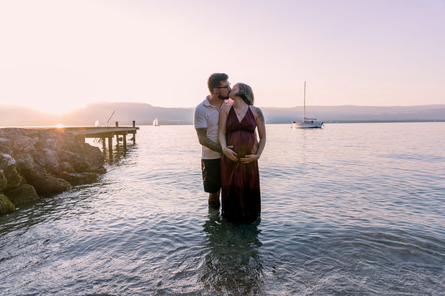 Pregnant couple kissing romantically while standing in calm lake water at golden sunset.
