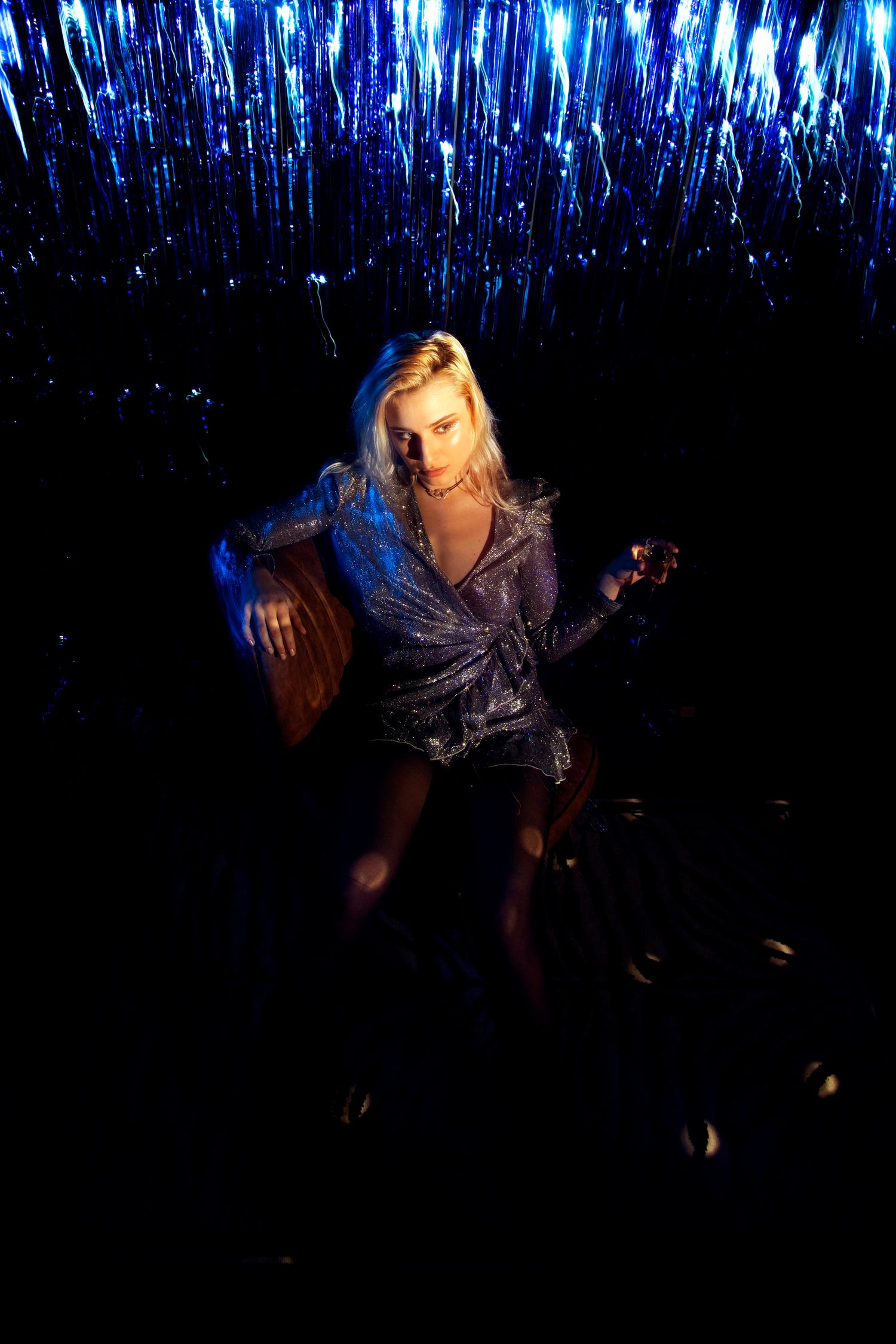 Blonde woman in silver mini dress poses in chair with blue sparkling light curtain backdrop.