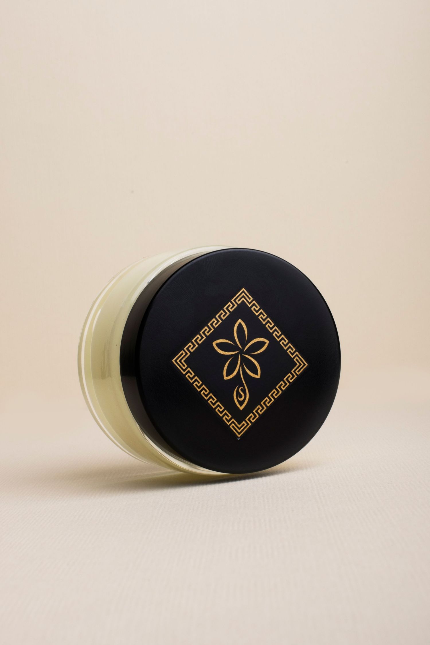 Luxury black and gold cosmetic jar with floral diamond logo on beige background.