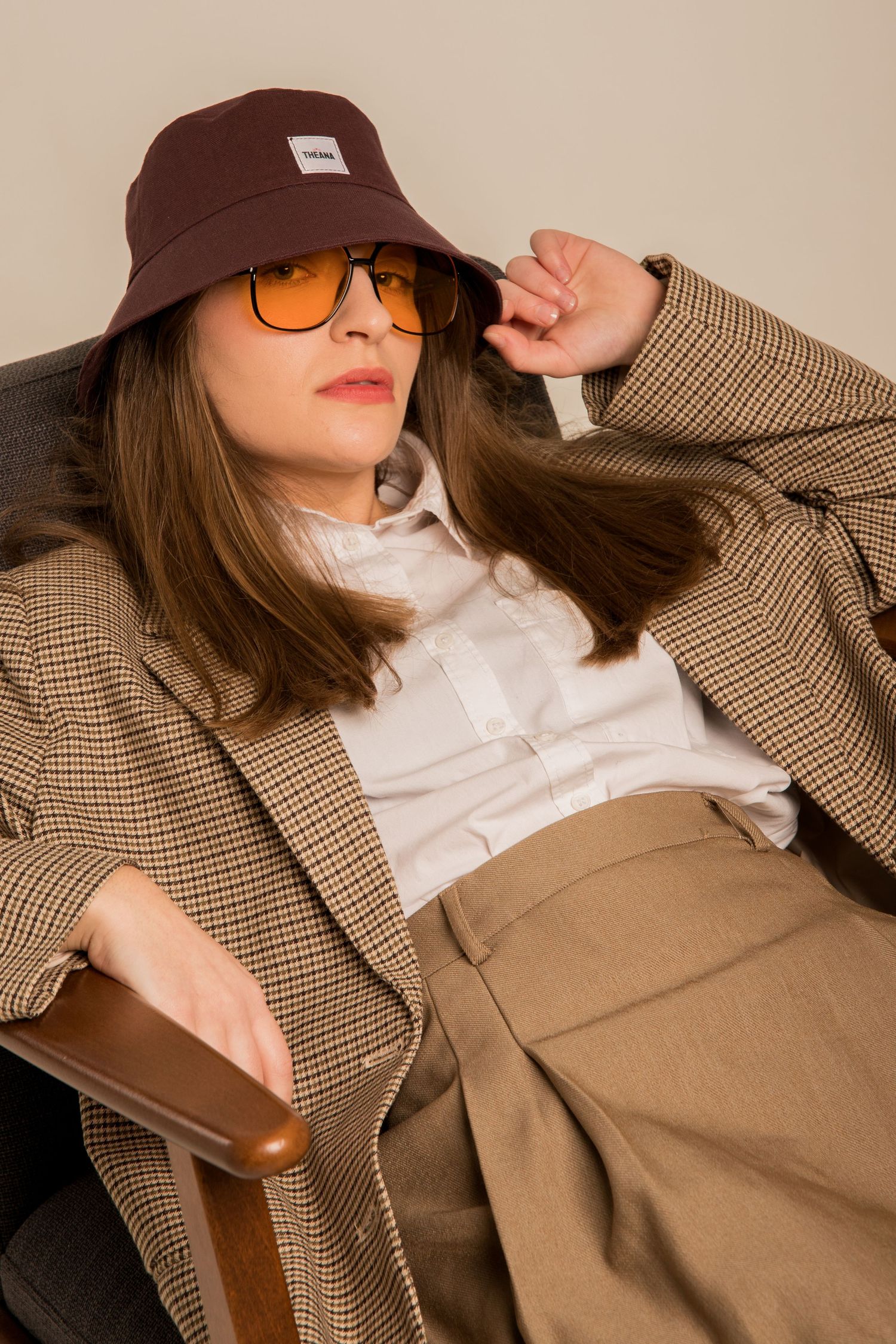 Woman in burgundy bucket hat and orange sunglasses relaxing in a chair wearing a plaid blazer.
