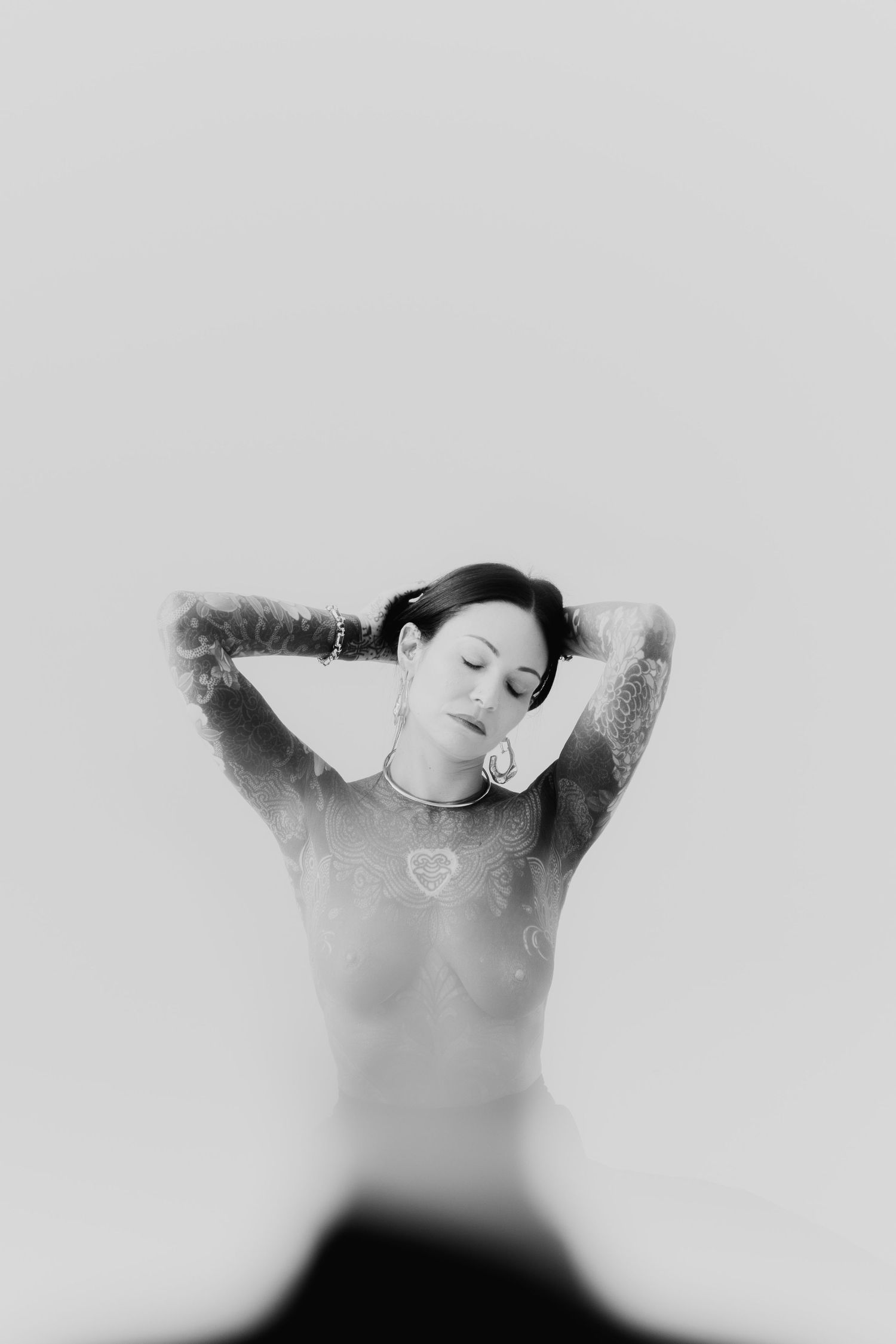 Black and white fine art portrait of tattooed woman with closed eyes and hands behind her head.