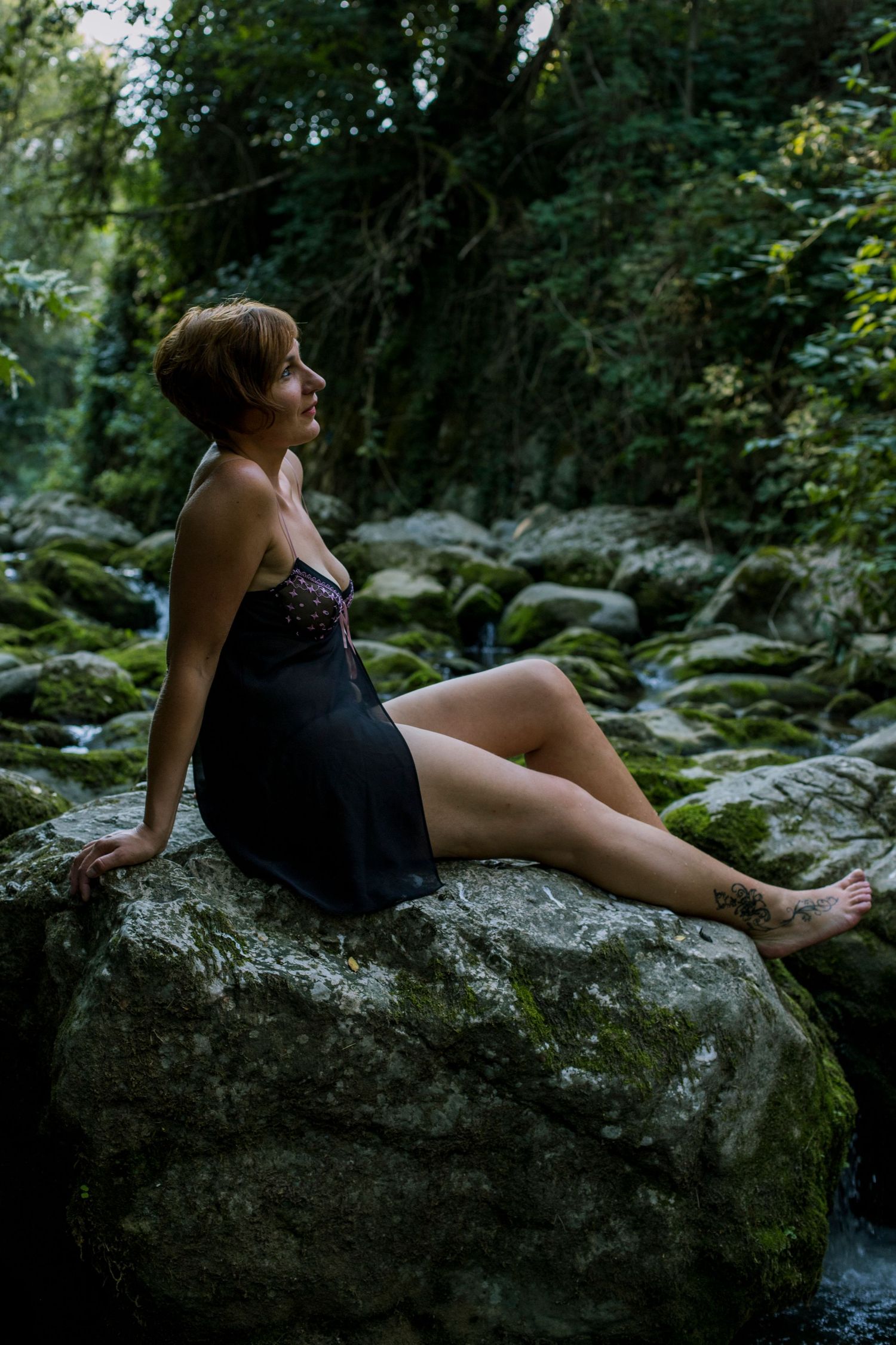 Woman in black slip dress sitting on mossy rocks in a lush forest setting.