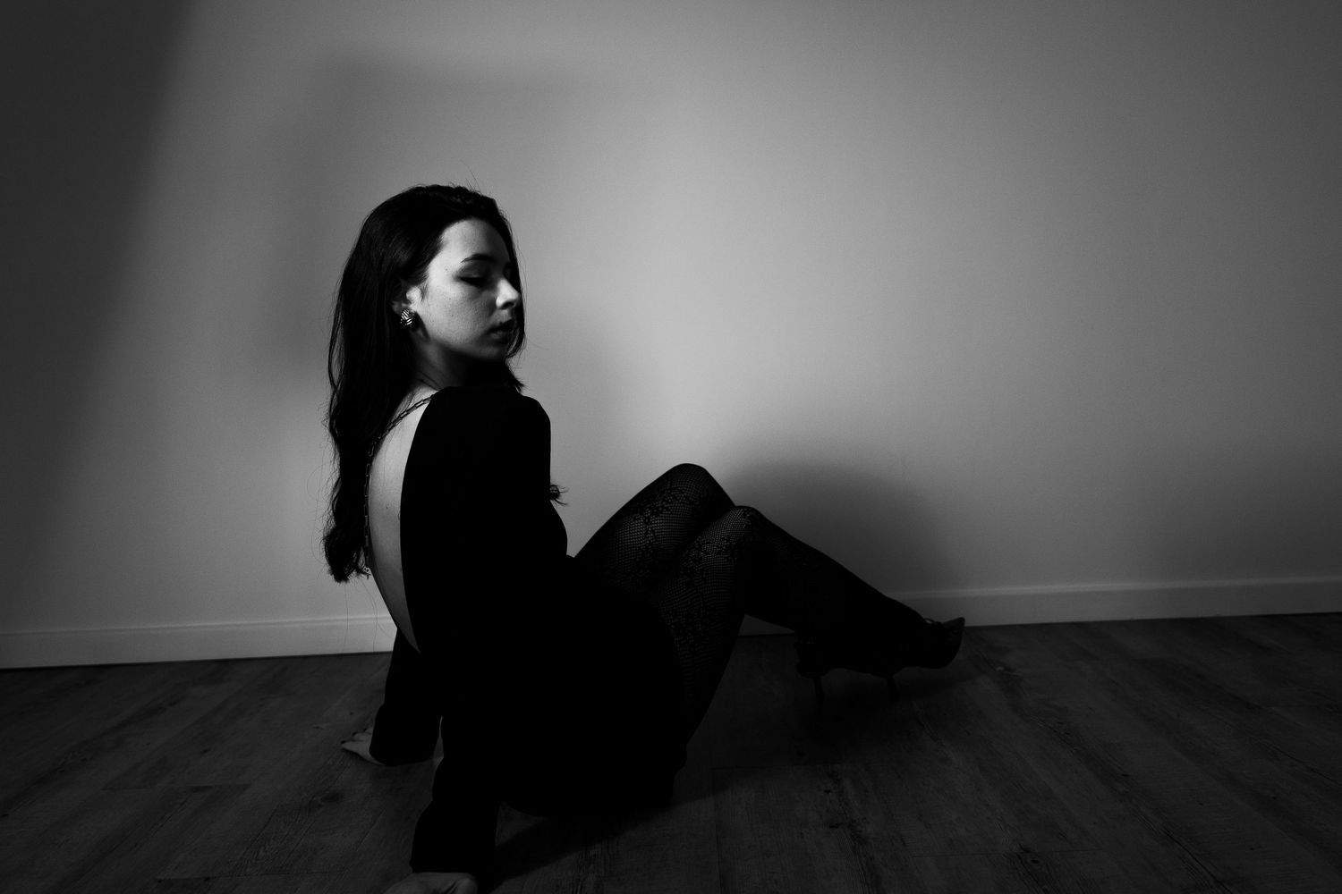 Black and white portrait of a woman with long dark hair sitting on a wooden floor against a white wall.