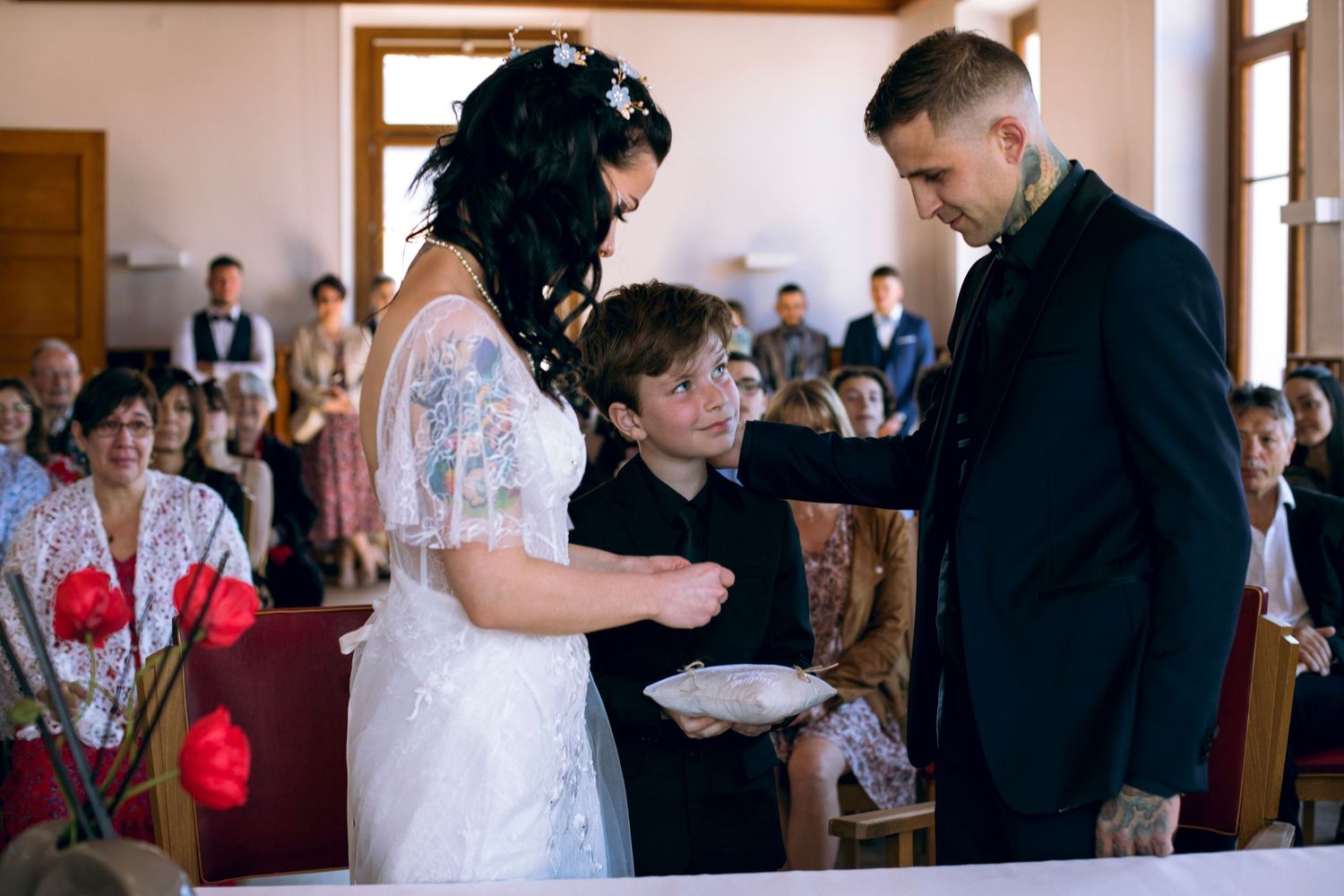 Bride and tattooed groom receive wedding rings from smiling young boy during indoor ceremony.
