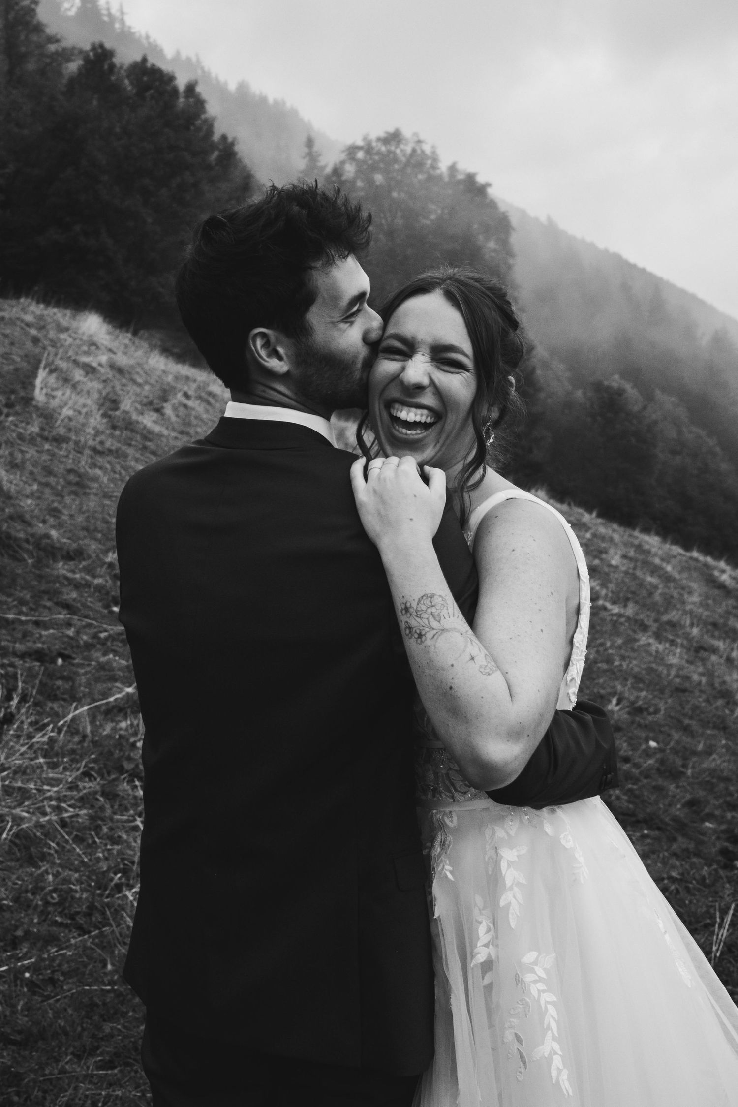 Black and white photo of groom kissing brides cheek as she laughs joyfully on a foggy mountain hillside.