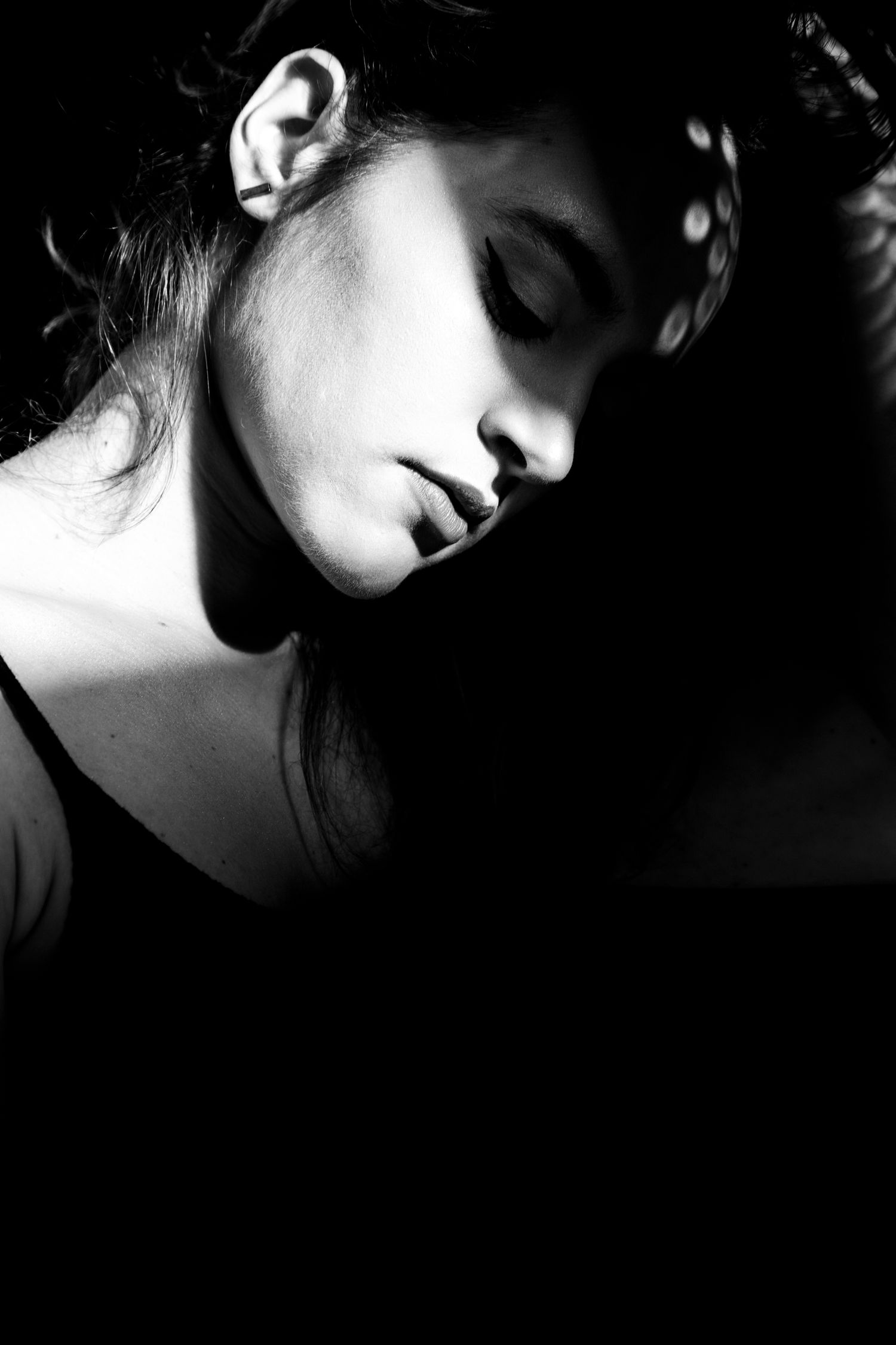 Black and white artistic portrait of a young woman with eyes closed, head tilted down in soft light.
