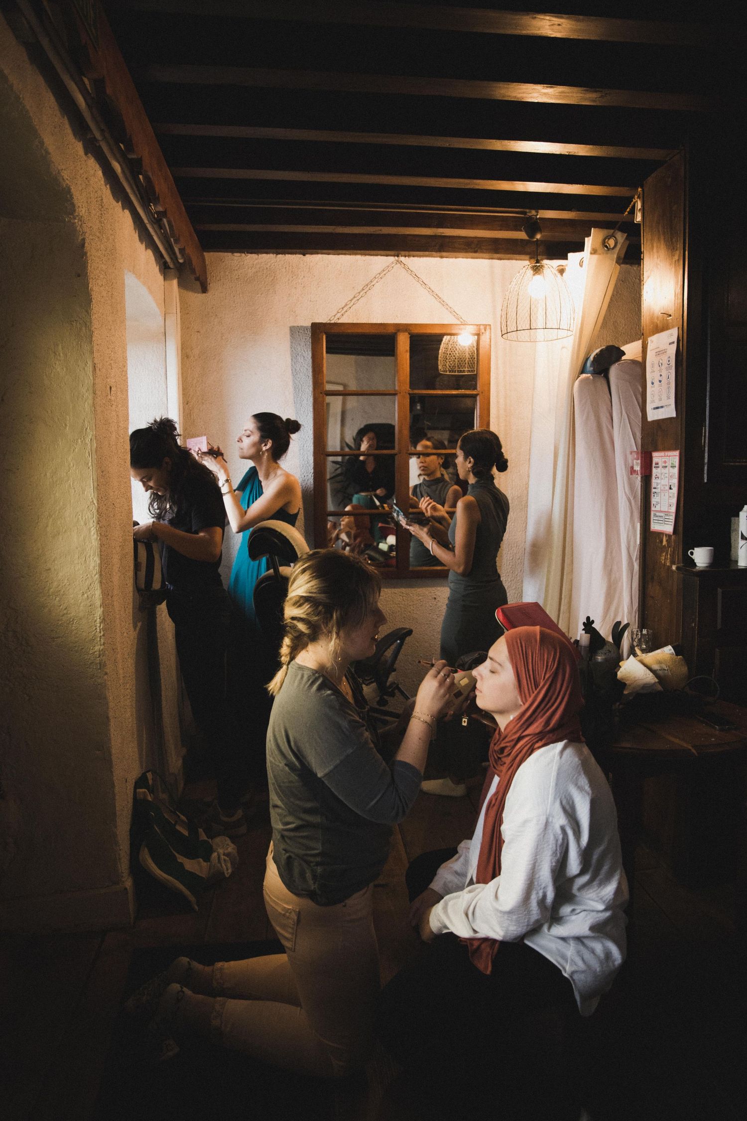 Group of women getting ready in a warmly lit rustic room, applying makeup and styling hair before an event.
