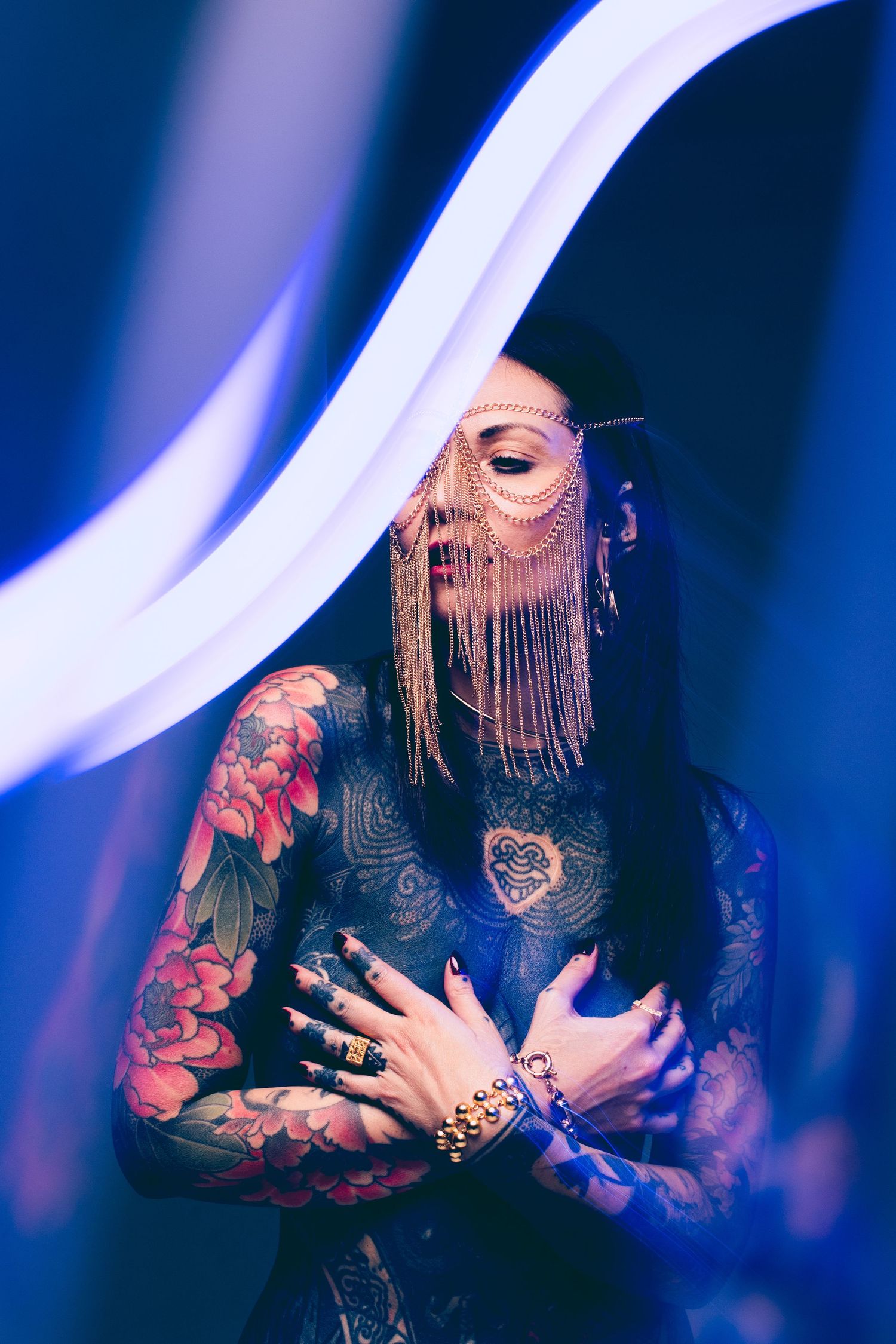 Tattooed woman in lace bodysuit with gold fringe face mask poses in blue neon light.