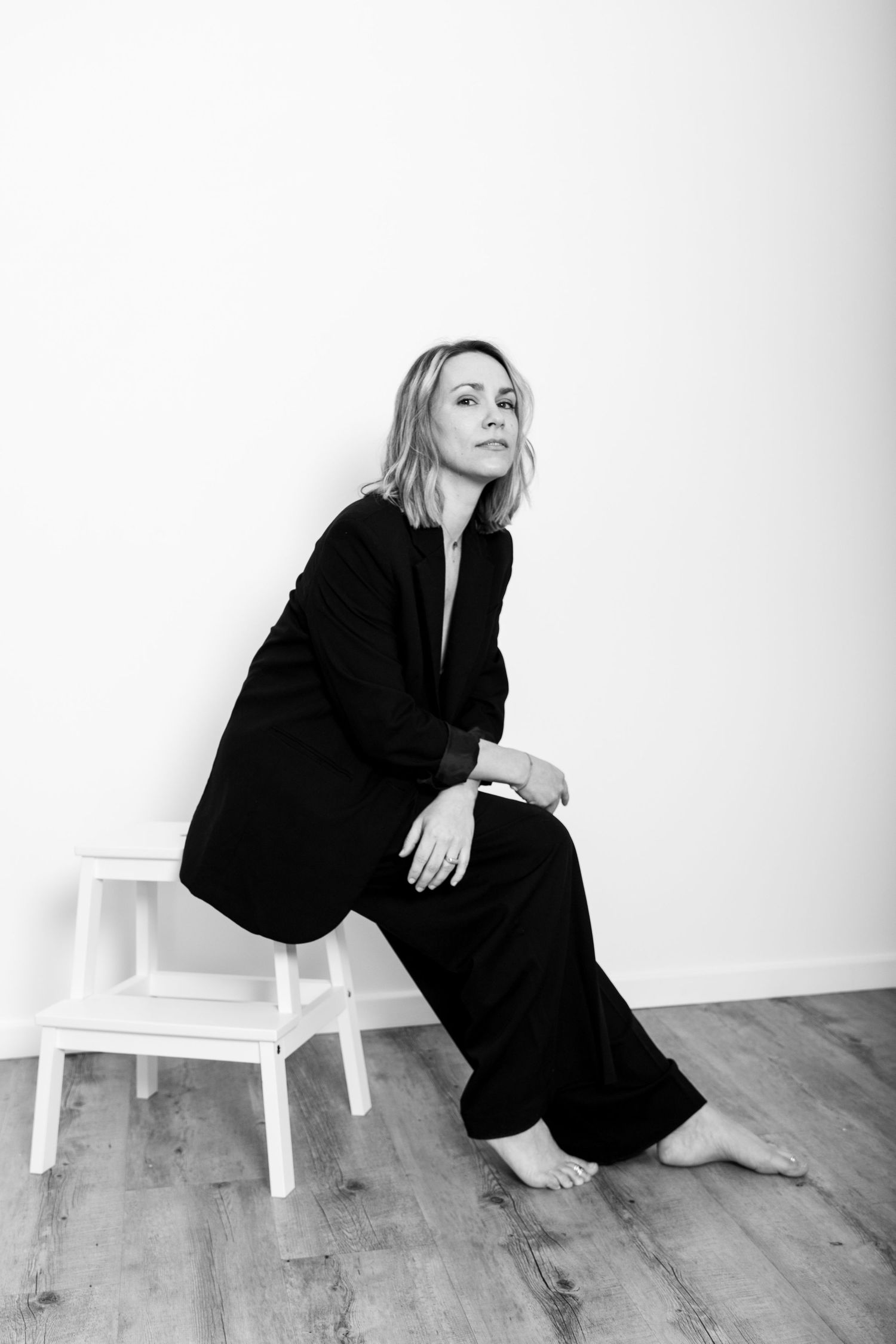 Woman in black suit sitting on white ladder stool, elegant black and white portrait.