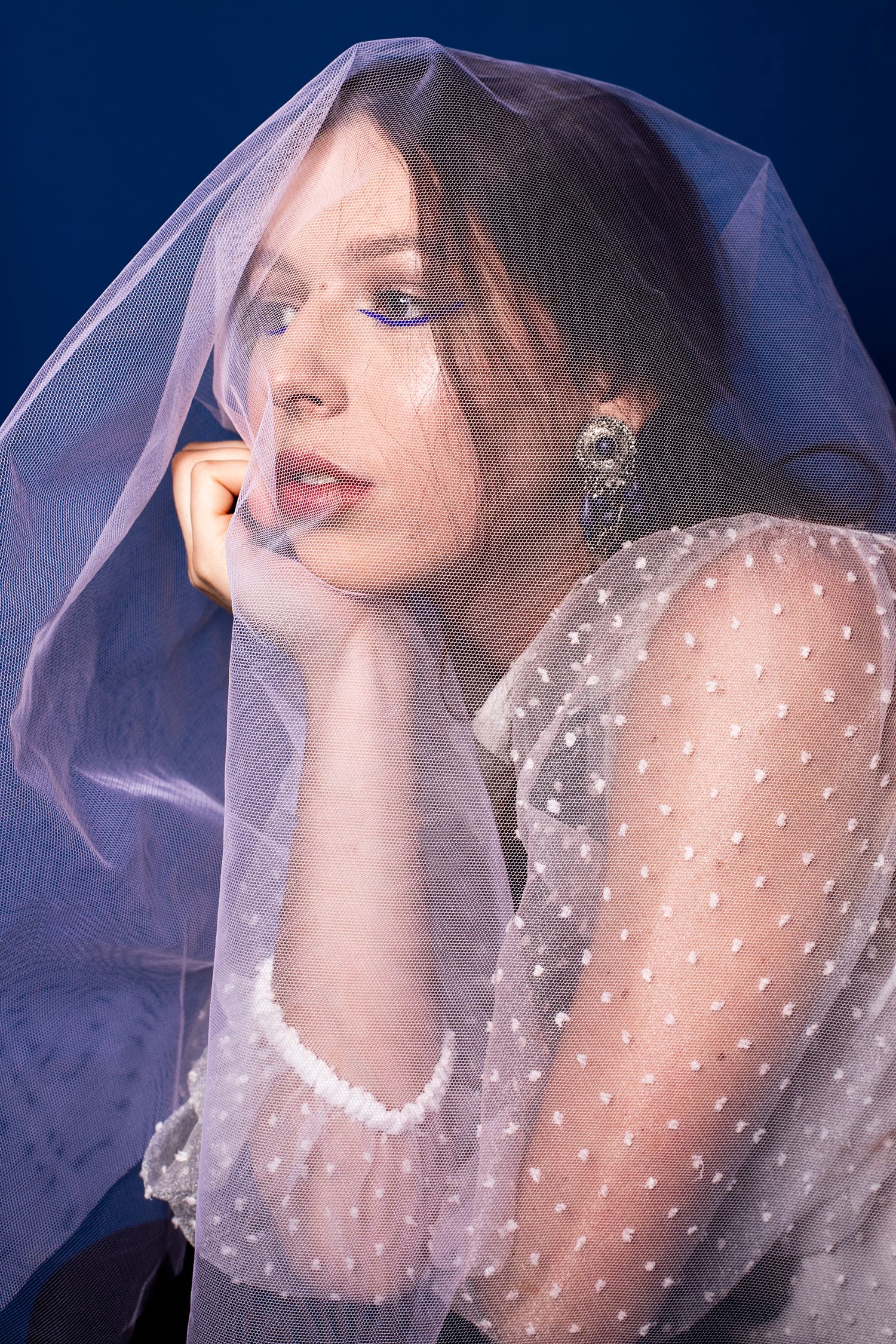 Woman draped in a sheer blue veil with dramatic eye makeup posing against a blue background.