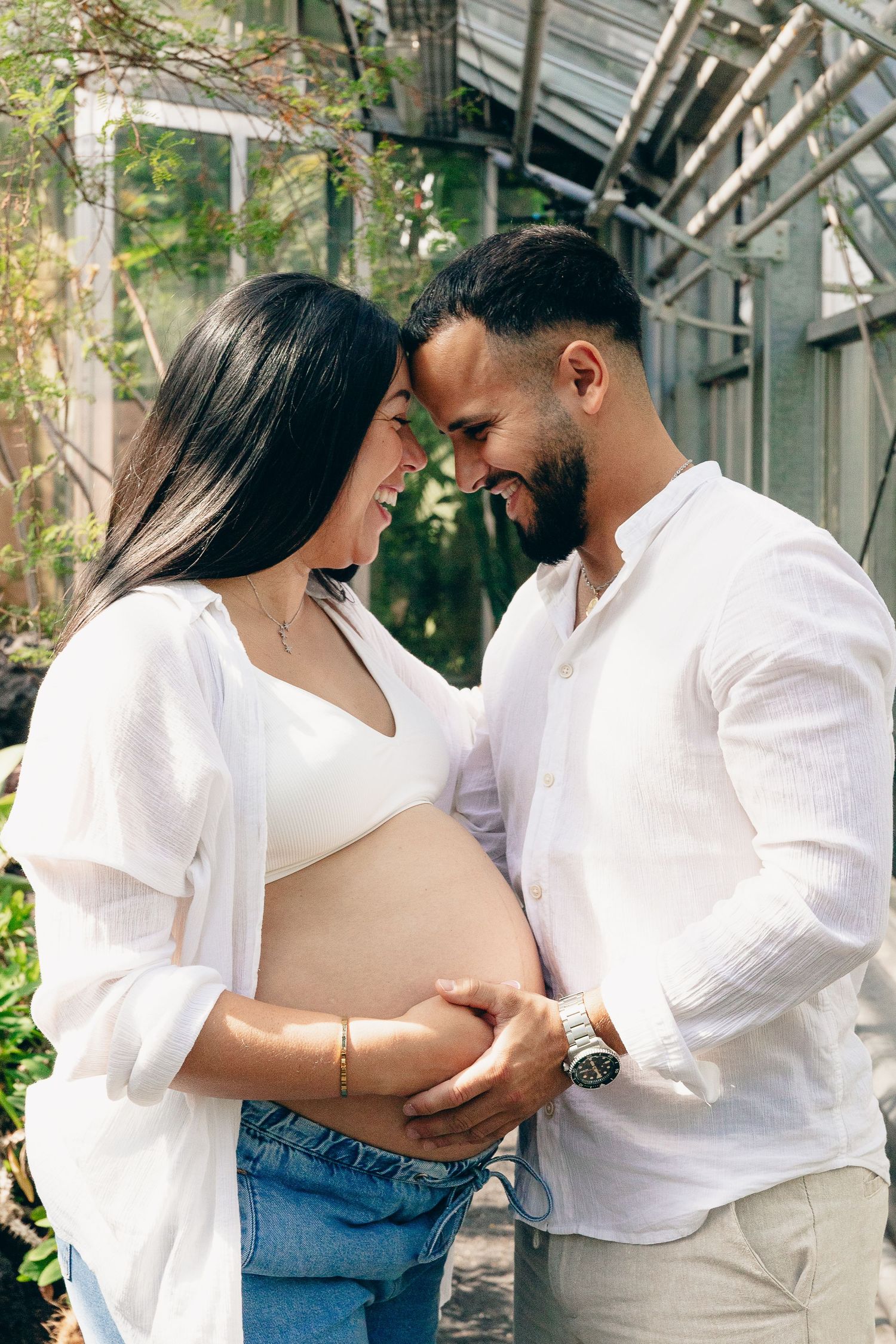 Happy couple smiling at each other while holding pregnant belly during outdoor greenhouse maternity session.