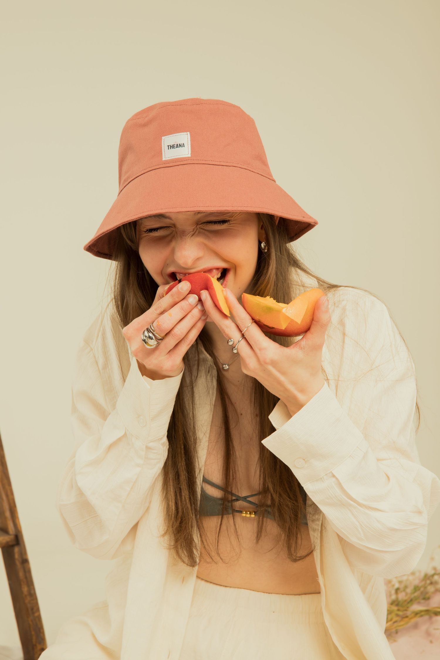 Woman in pink bucket hat biting into fresh melon slice, wearing white outfit in neutral studio setting.