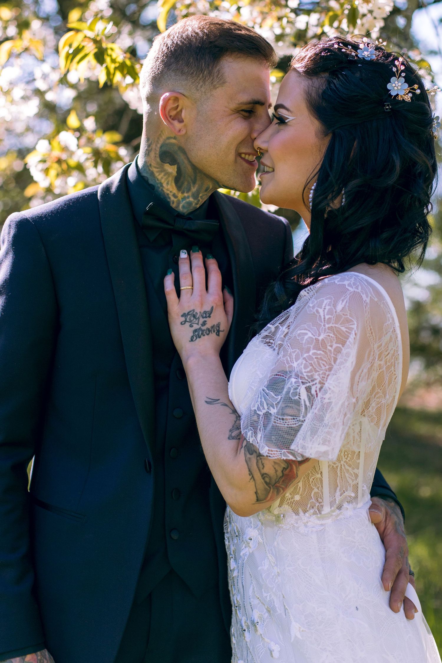 Tattooed bride and groom share a close romantic moment near blooming spring trees.