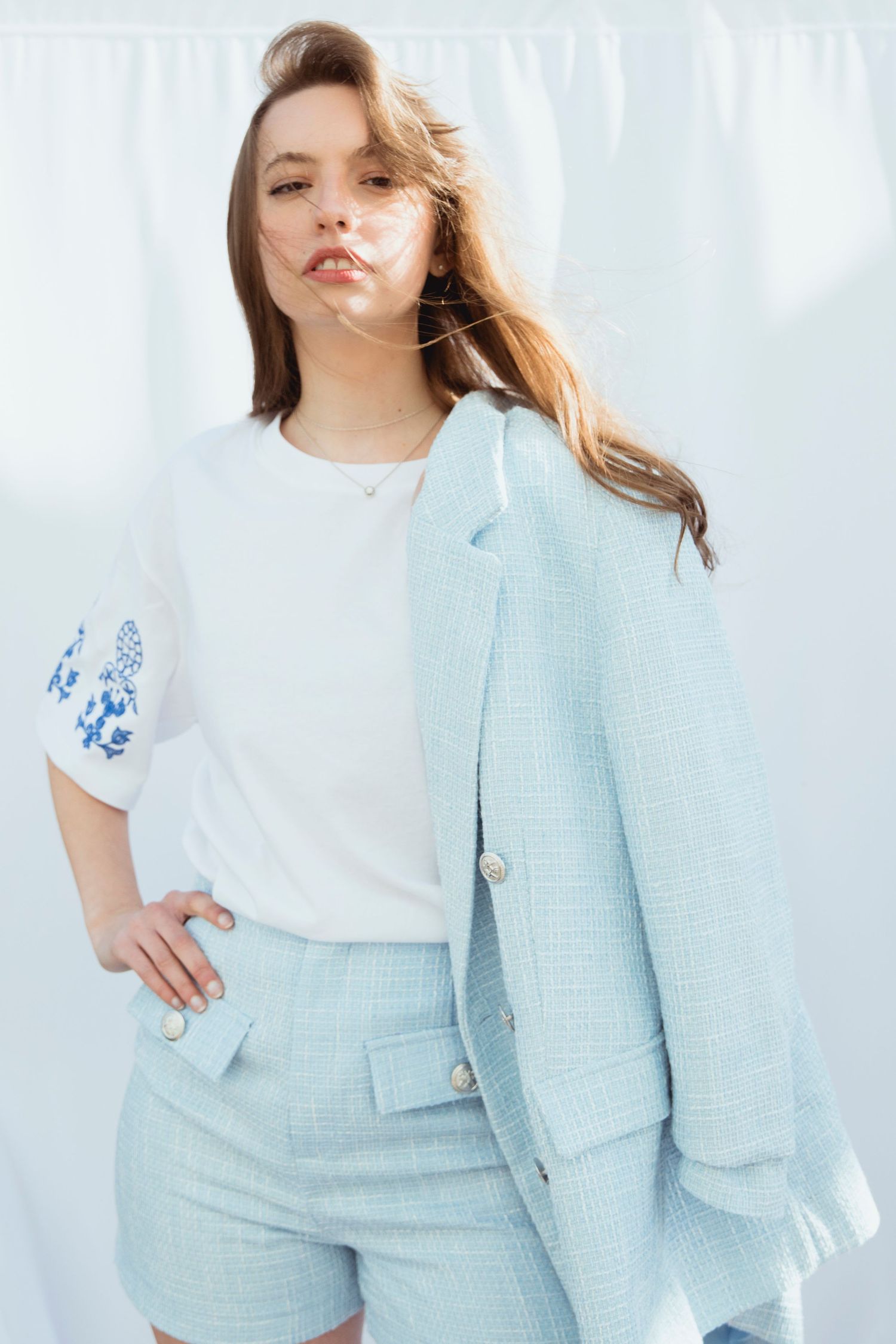 Woman in a white embroidered top and light blue blazer posing outdoors with wind-blown hair.