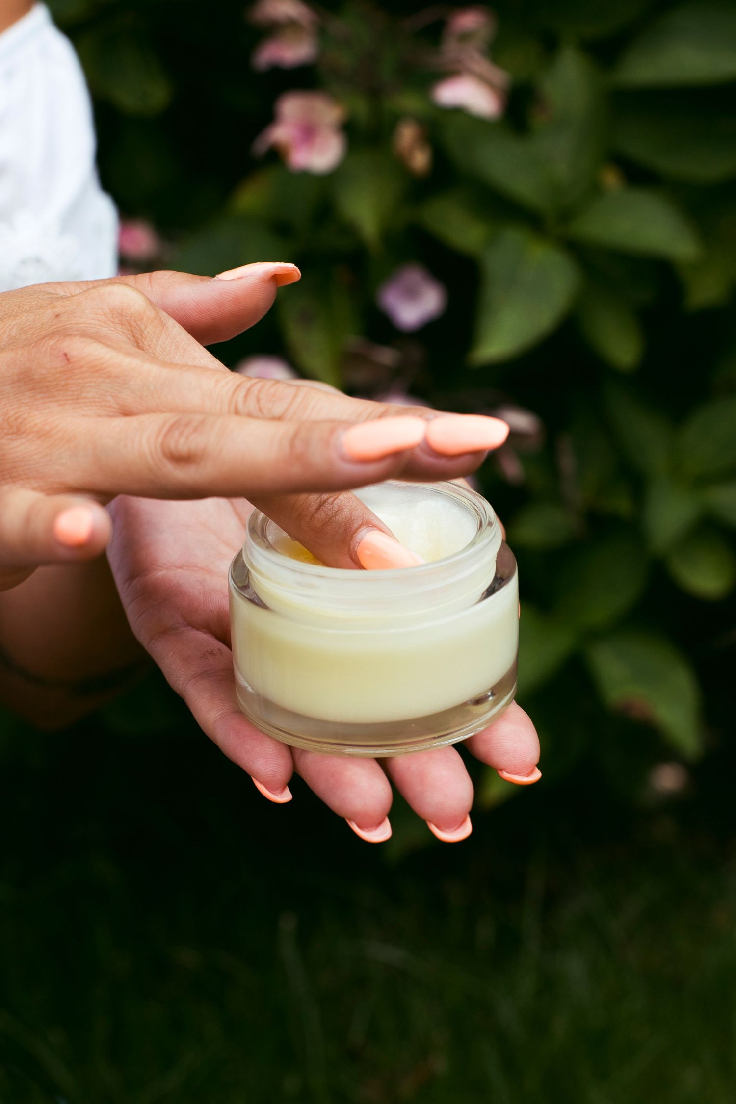 Woman applies white cream from open glass jar outdoors with green floral background.