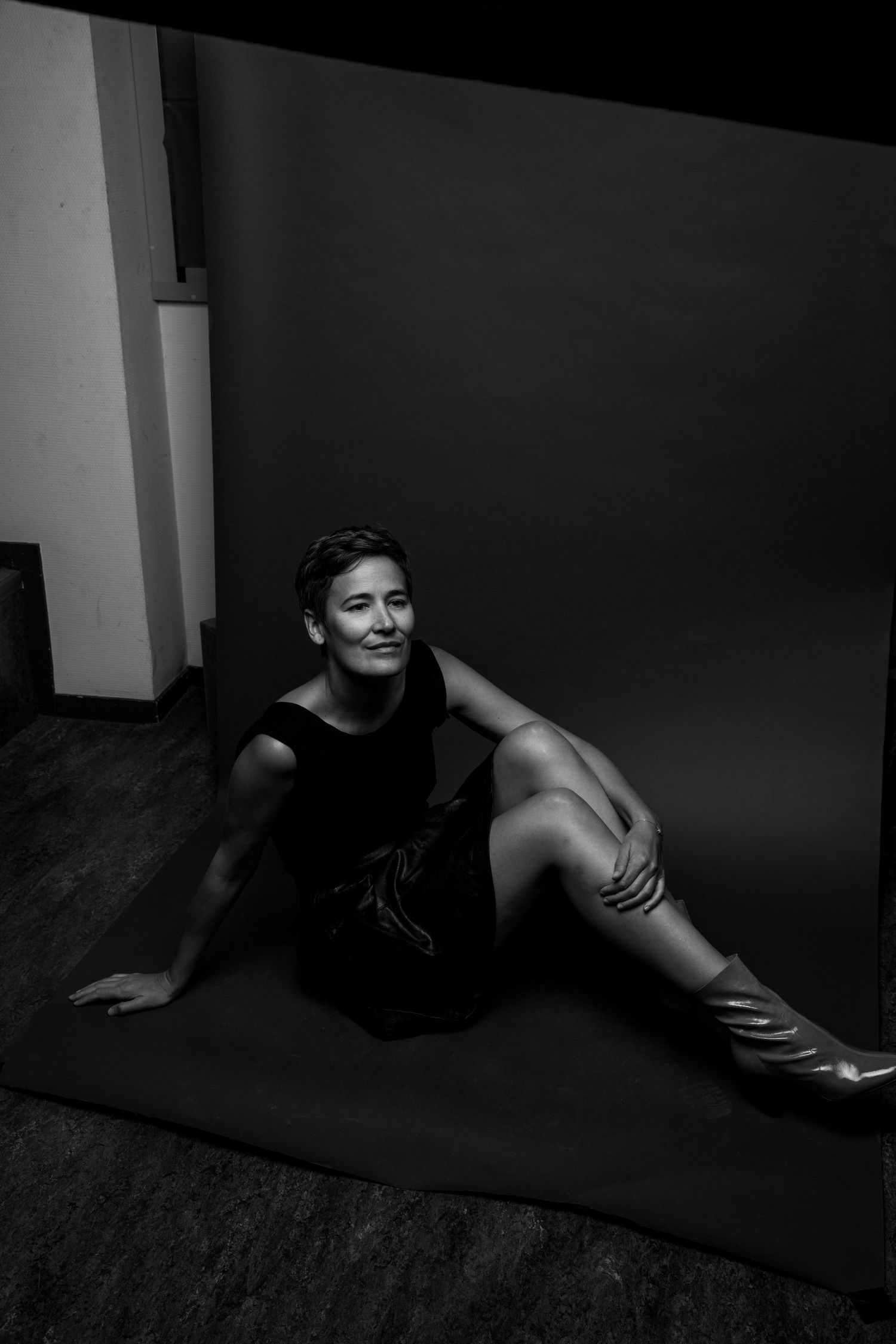 Woman in black dress sitting gracefully on floor against dark backdrop in black and white studio portrait.