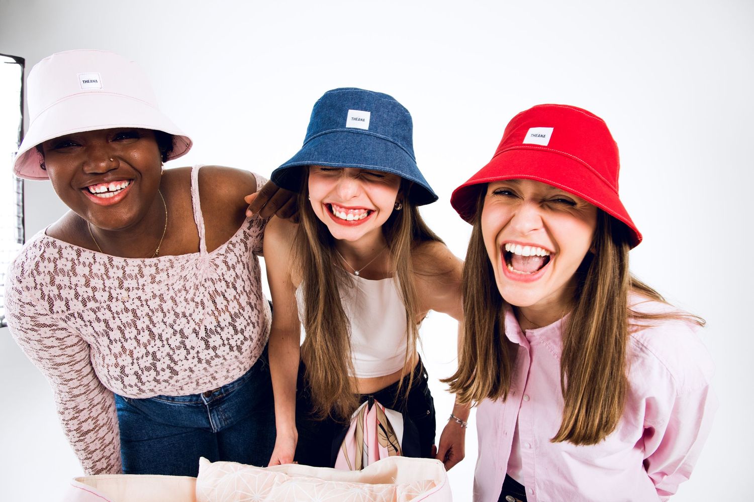 Three laughing young women wearing colorful bucket hats pose together against a white background.