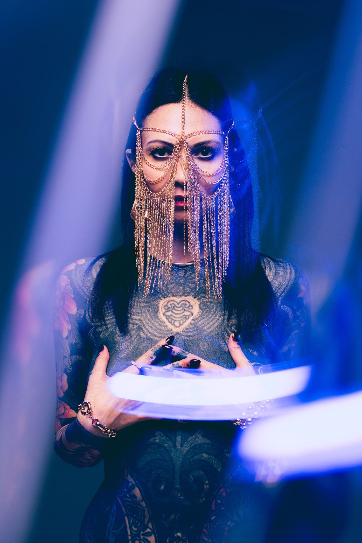 Tattooed woman wearing an ornate gold chain face veil holds a glowing object in blue neon light.