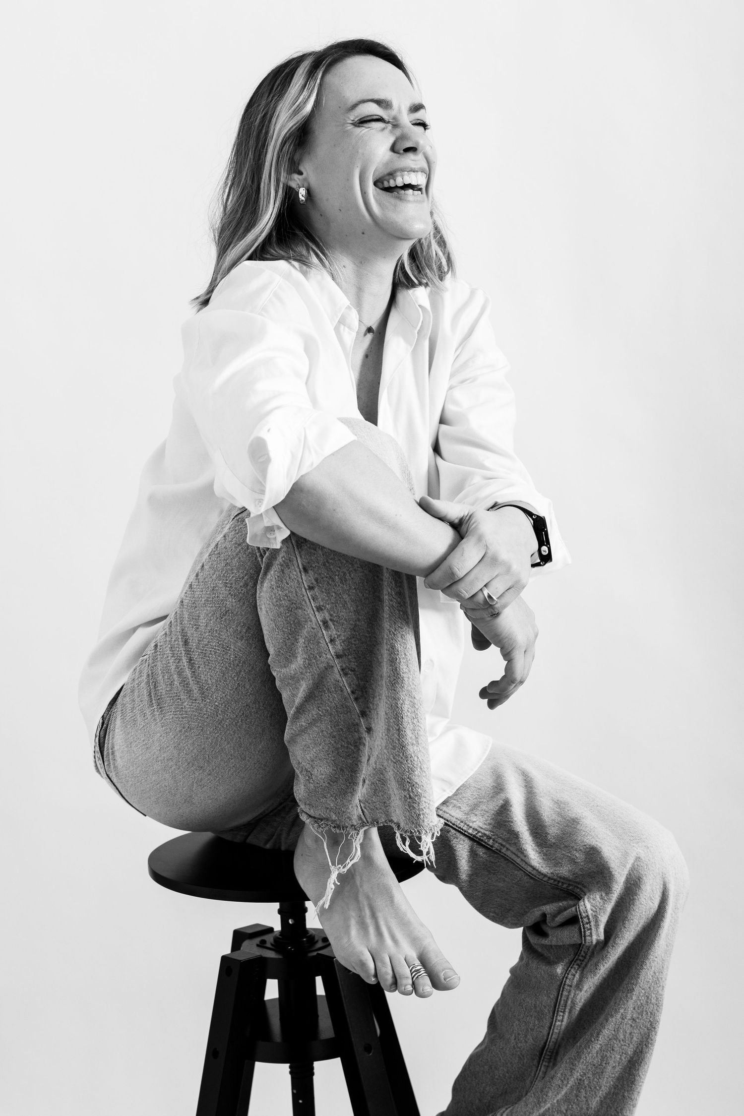 Black and white photo of laughing blonde woman sitting on stool in white shirt and jeans.