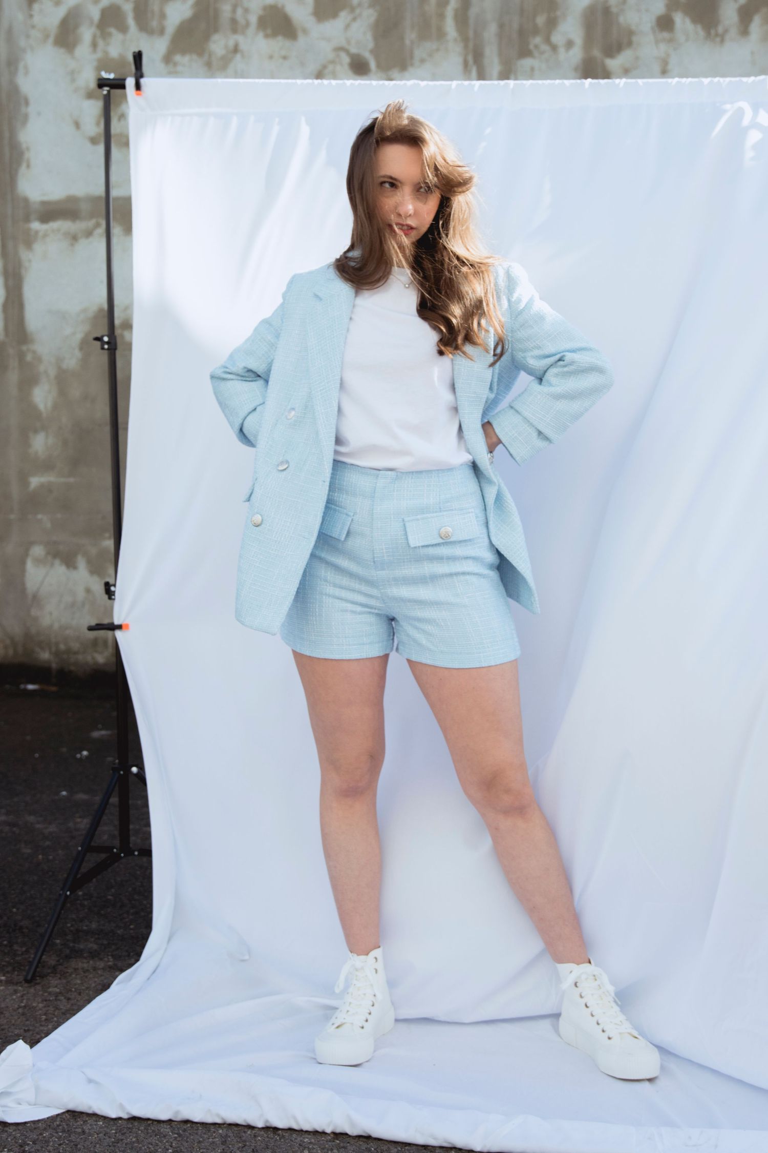 Young woman posing in a light blue blazer, matching shorts, and white sneakers against a white backdrop.