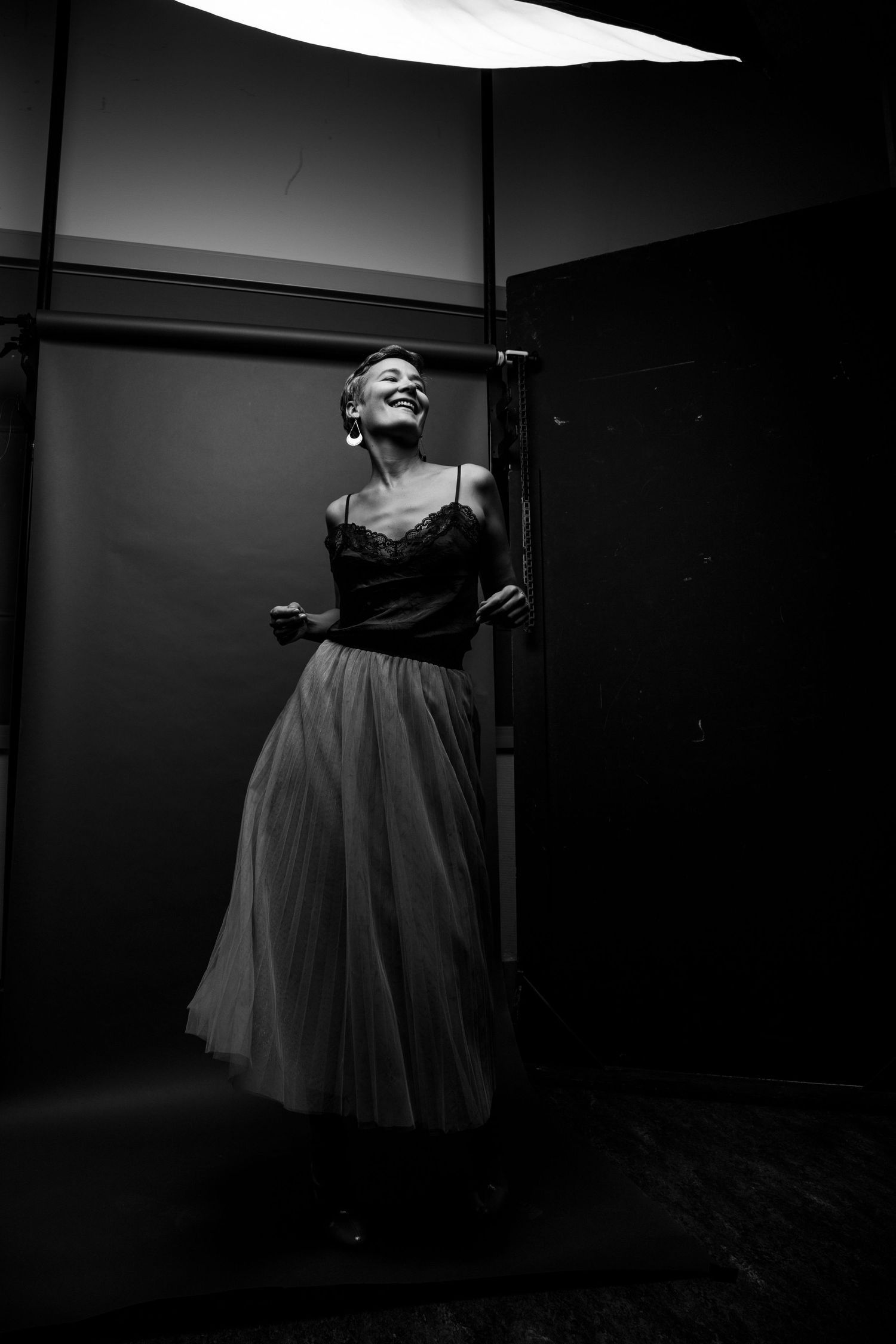 Woman in black lace top and white tulle skirt laughing joyfully under studio light in black and white photo.