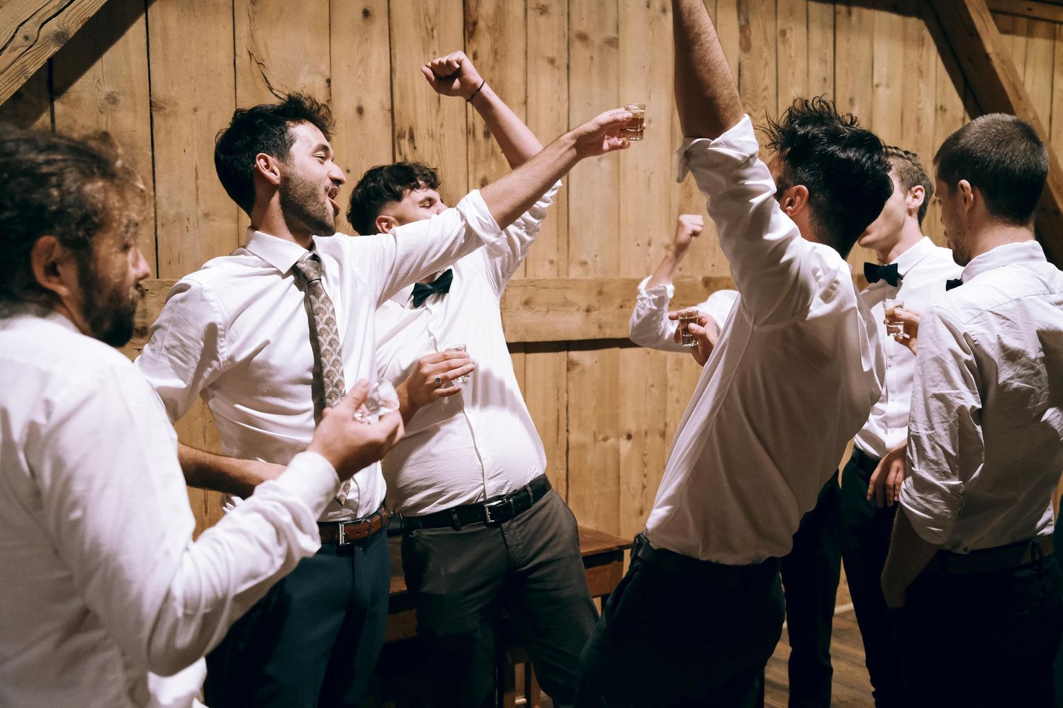 Group of young men in white shirts dancing and celebrating energetically inside a rustic wooden barn.
