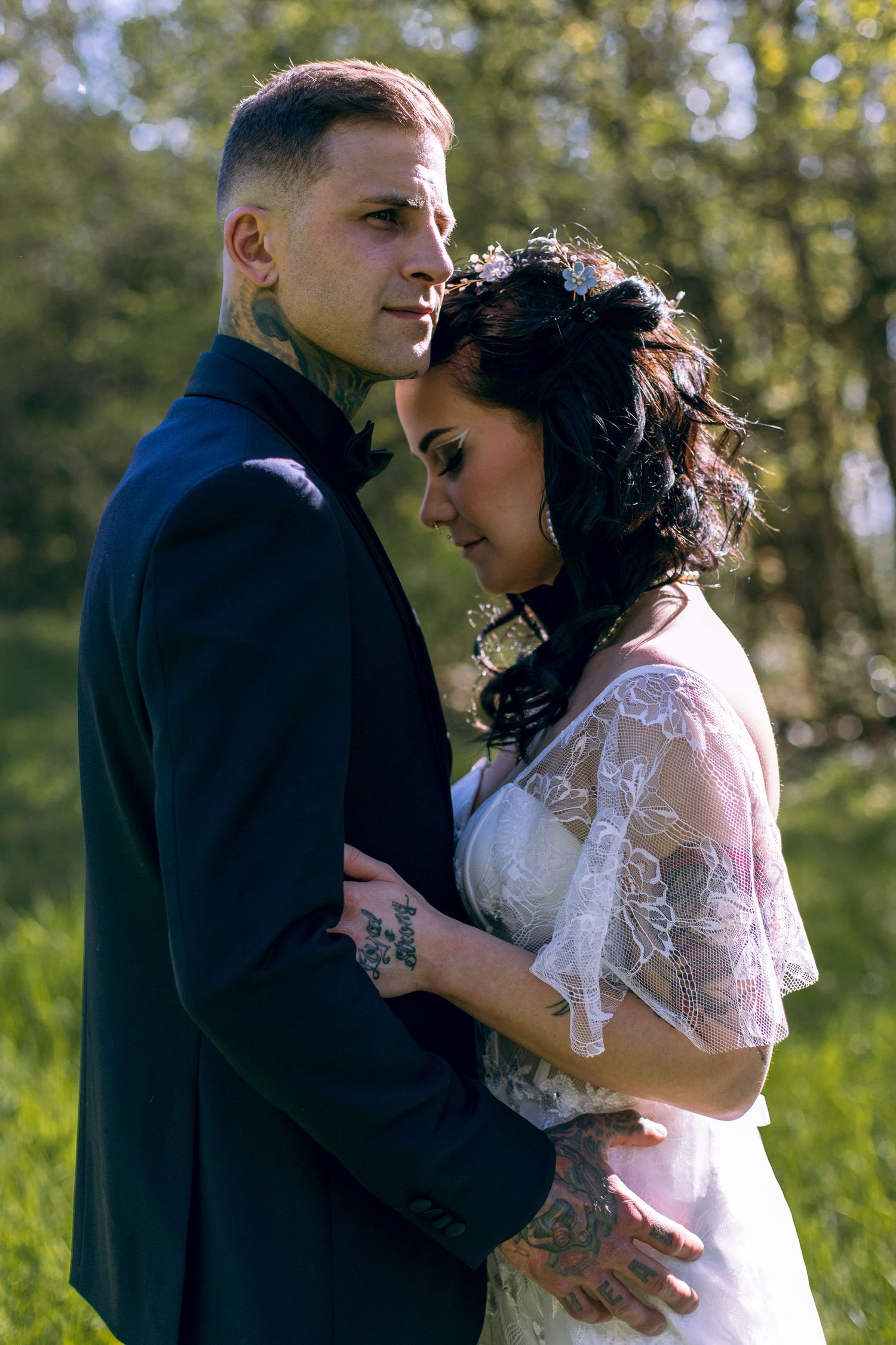 Tattooed bride and groom embrace tenderly in romantic outdoor nature setting.
