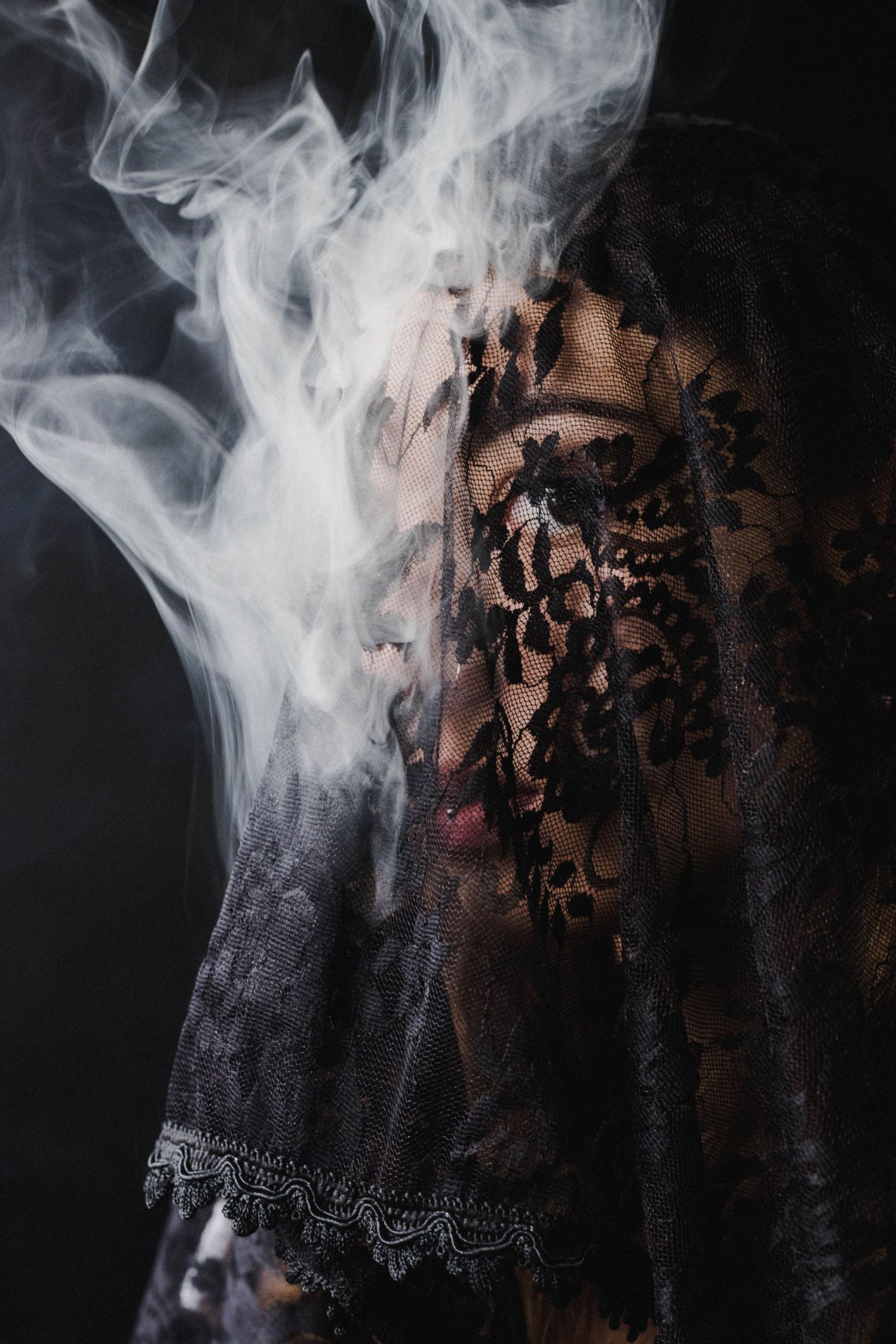Mysterious figure covered in black floral lace fabric with swirling white smoke against dark background.