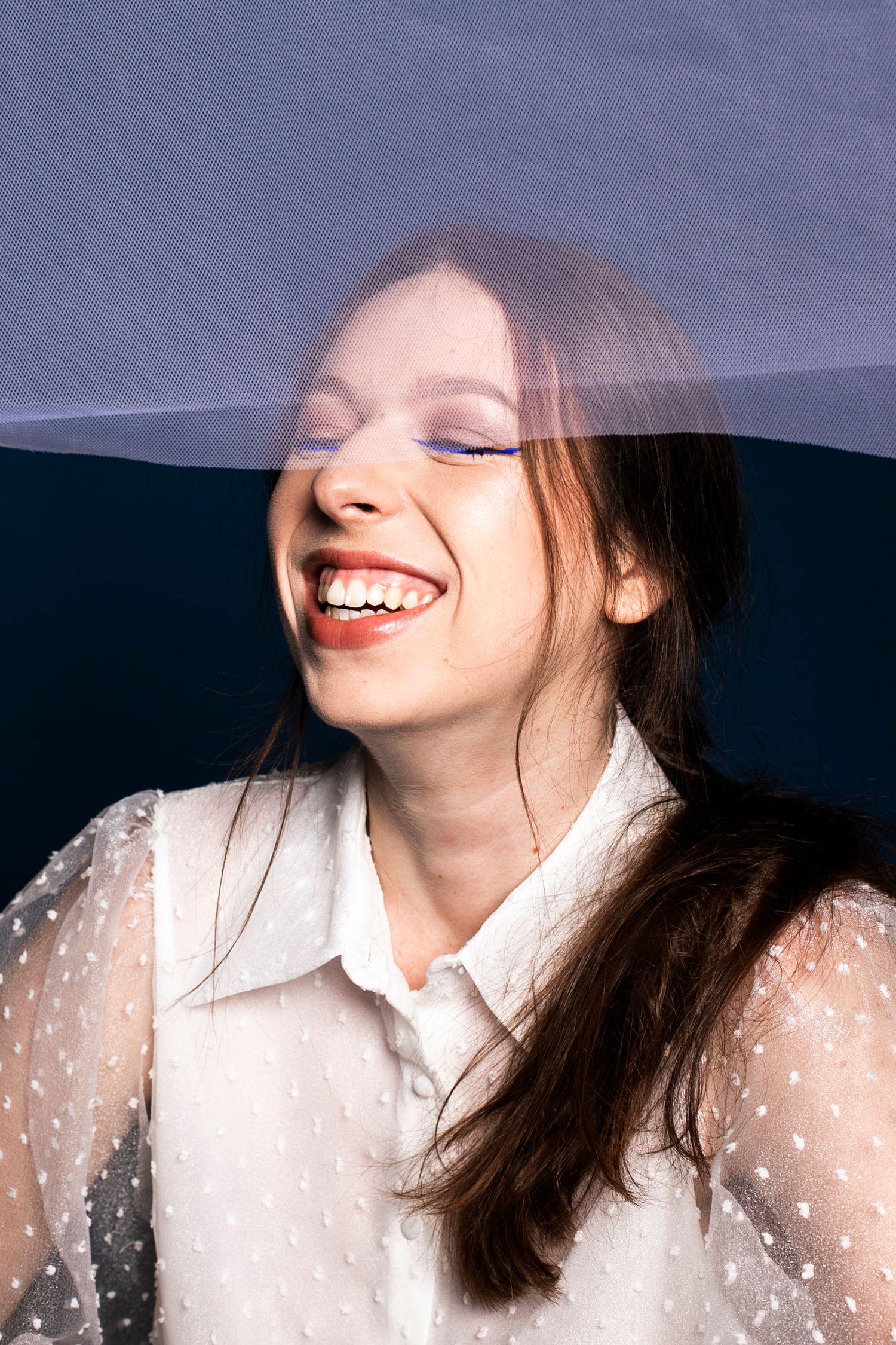 Laughing woman with closed eyes and red lips under a translucent white veil in a white sheer top.