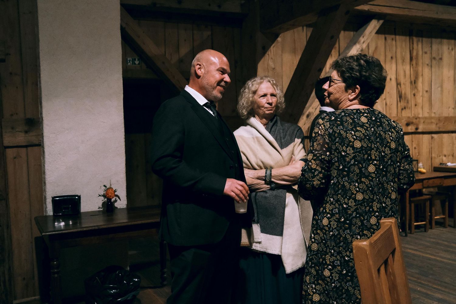 Three elegantly dressed wedding guests share a conversation inside a rustic wooden barn reception hall.