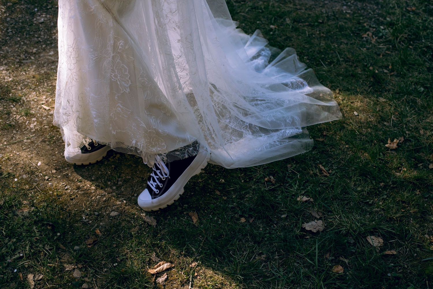 Close-up of bride wearing white lace wedding dress hem with black Converse sneakers on grass.