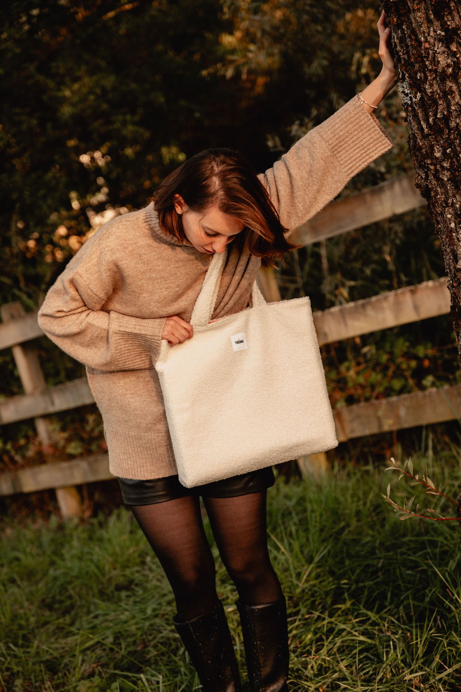 Woman in beige knit sweater looking into a cream textured tote bag while leaning against a tree.