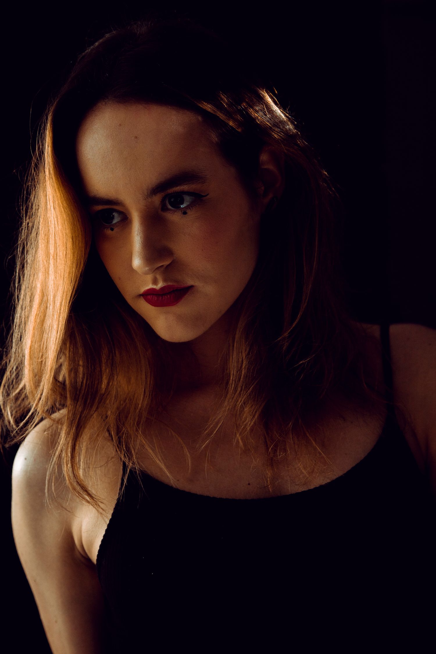 Dark dramatic portrait of a young woman with red lips and auburn hair in black tank top.