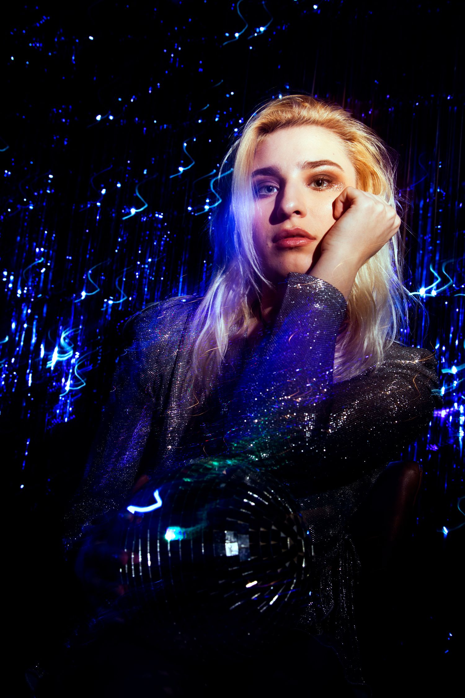 Blonde woman in sparkly jacket rests chin on hand while holding disco ball in blue light.