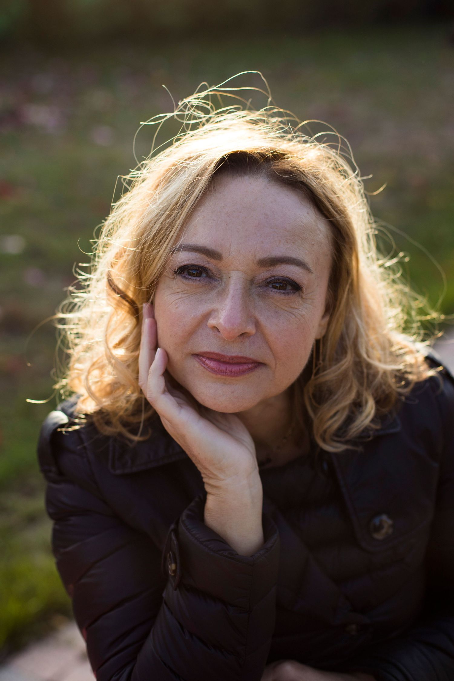 Portrait of a blonde woman resting her chin on her hand, bathed in warm sunlight in a park.