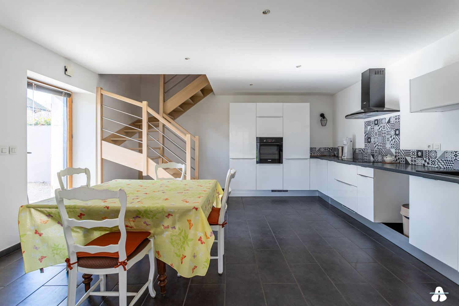 Modern open-plan kitchen with white cabinets, dark tile floors, dining table and staircase in a contemporary home.