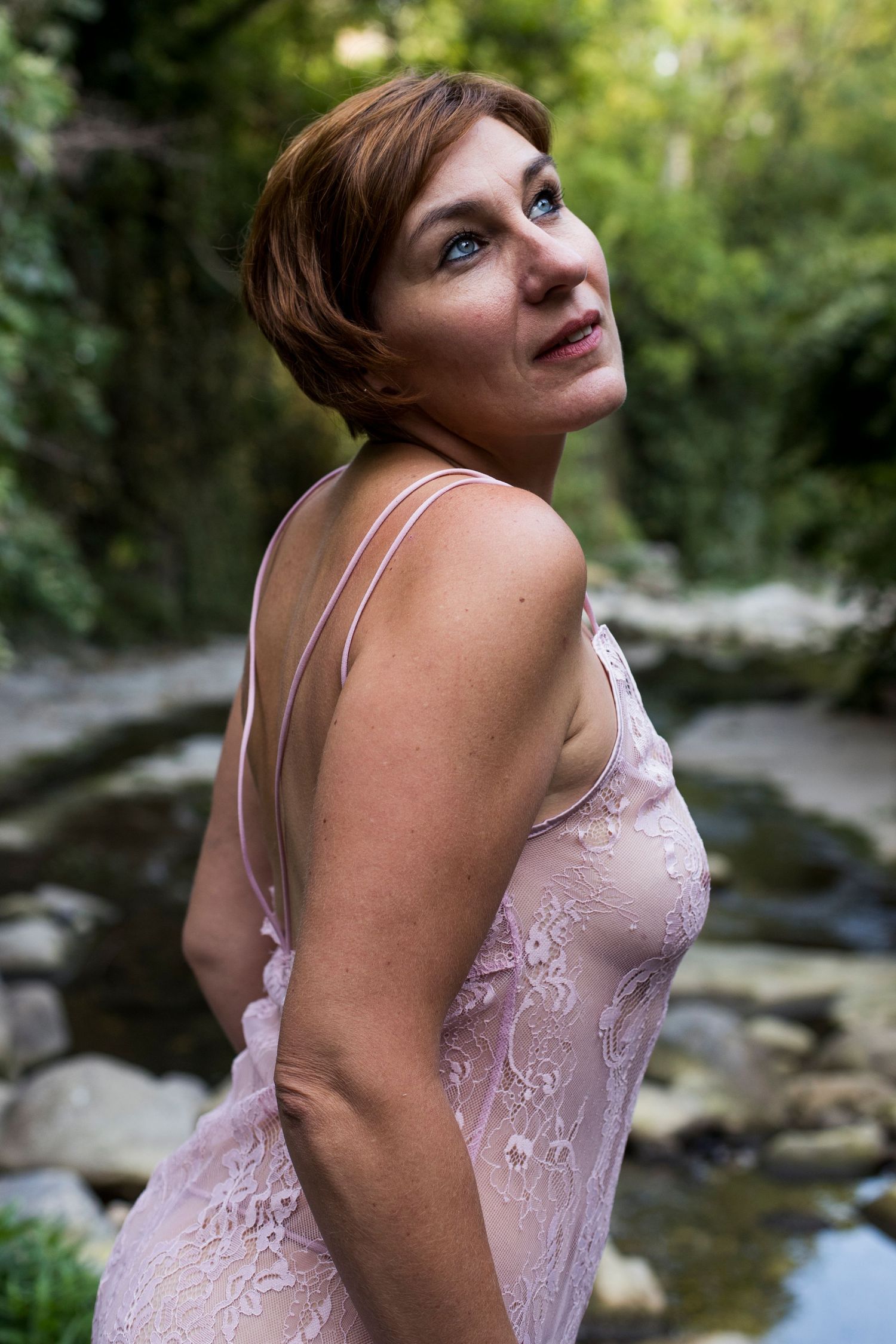 Woman in sheer pink lace dress standing near a forest stream.