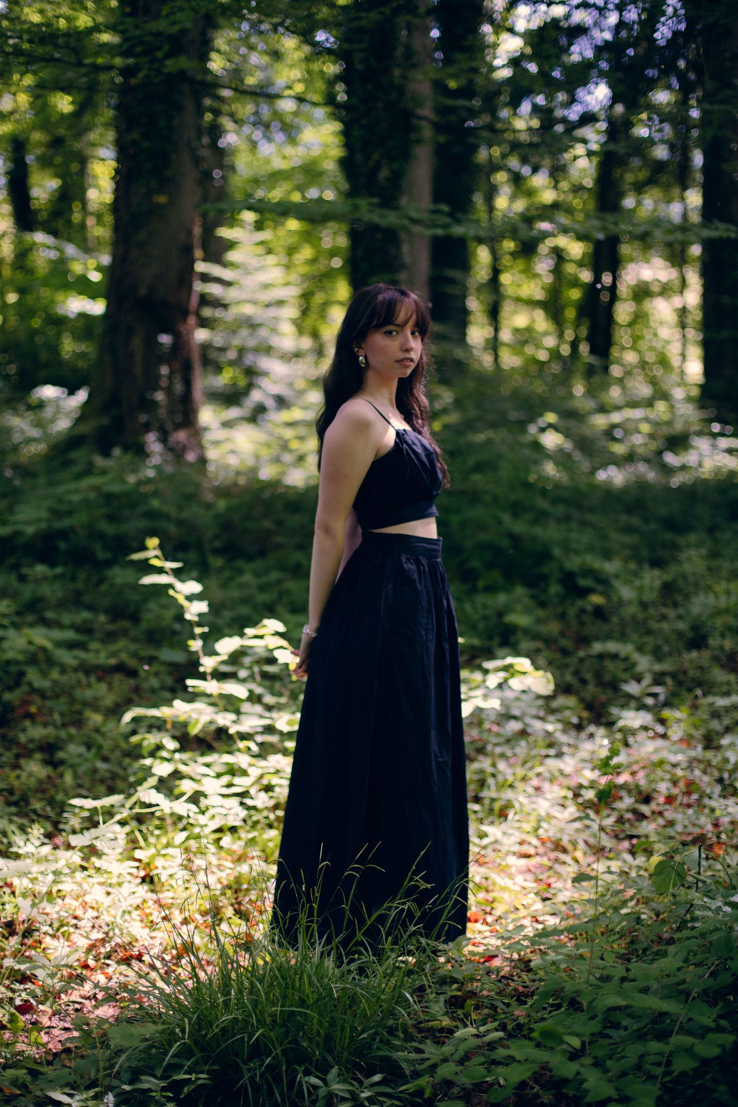 Young woman in black crop top and maxi skirt standing in sunlit forest.
