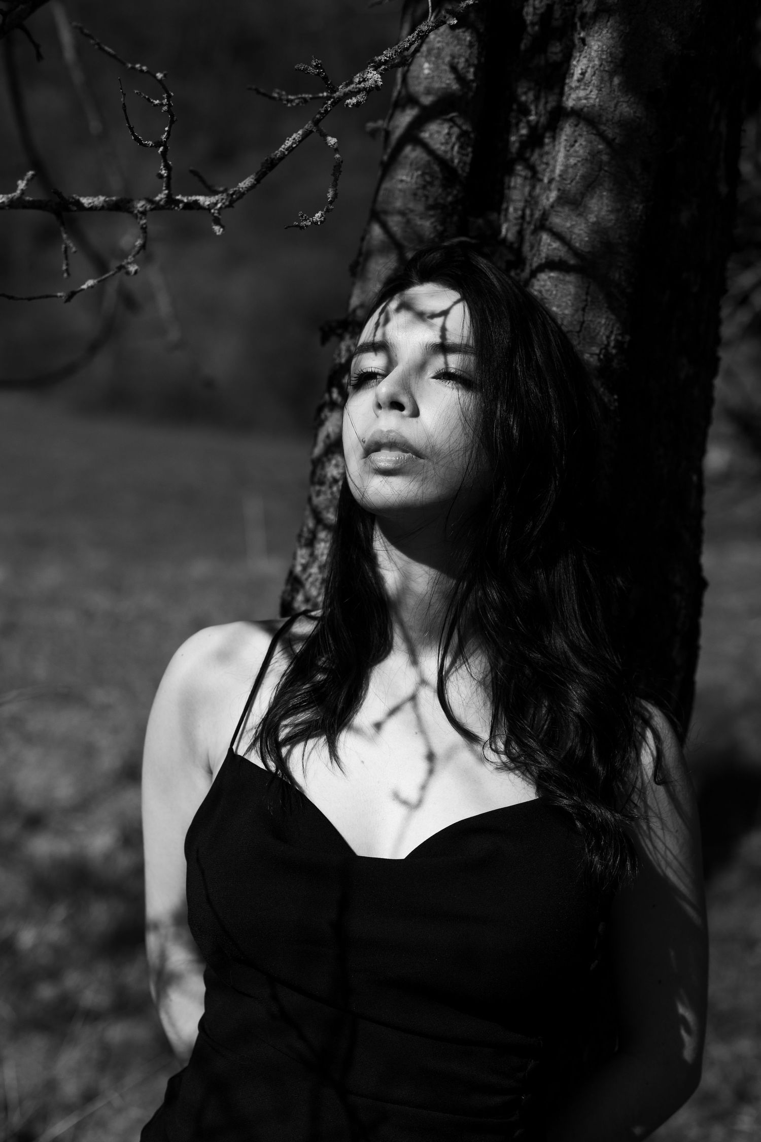 Black and white portrait of dark-haired woman leaning against tree with branch shadow patterns.