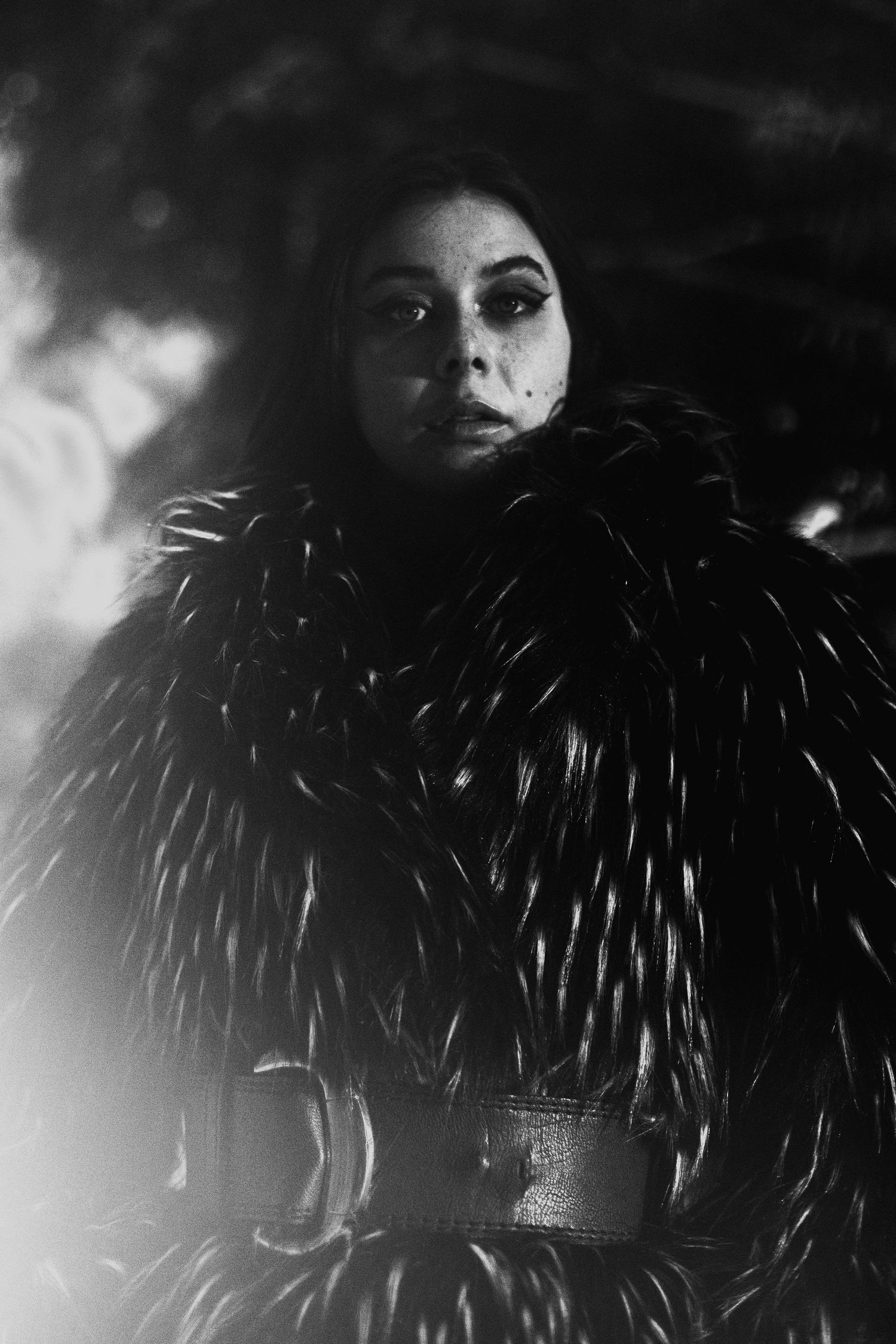 Dramatic black and white portrait of a woman in a fur coat with bold eye makeup in dark setting.