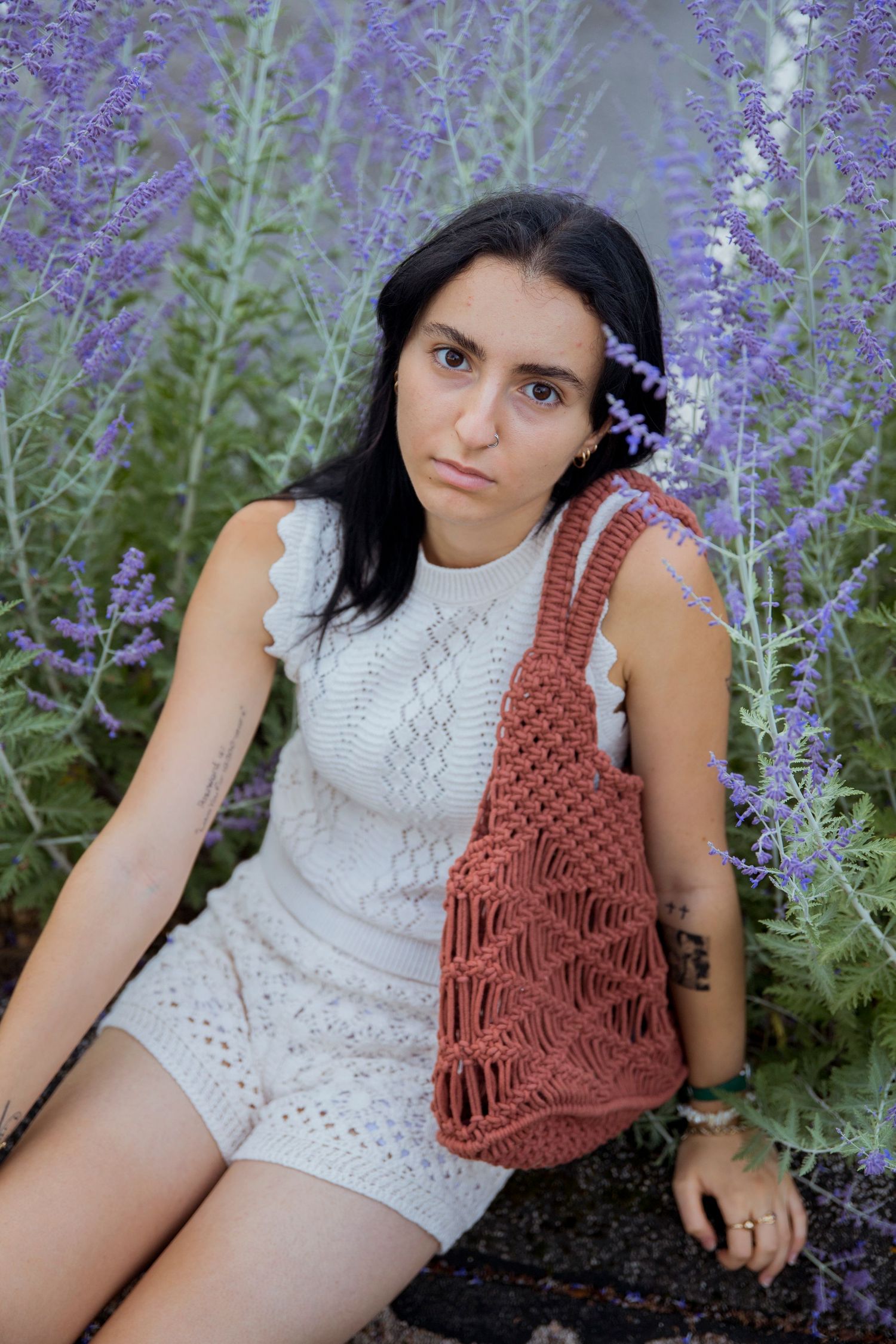 Young woman in white knit romper holding terracotta macrame bag among lavender flowers.