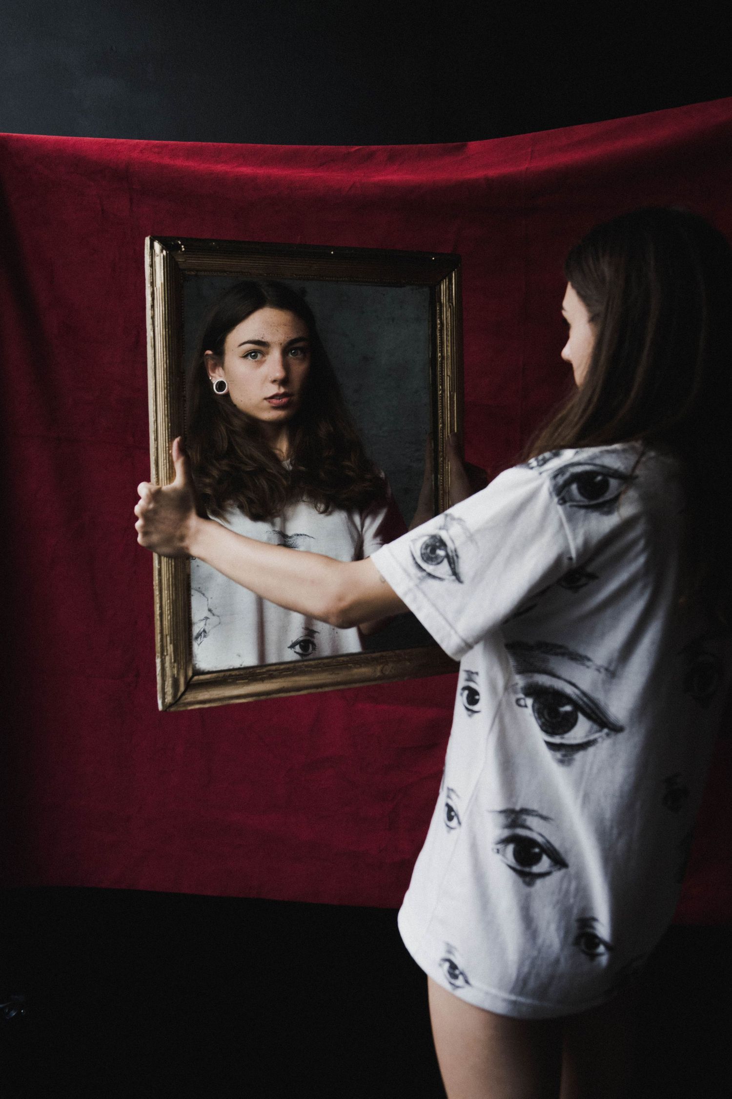 Young woman holding ornate mirror reflecting her image against dark red fabric backdrop.