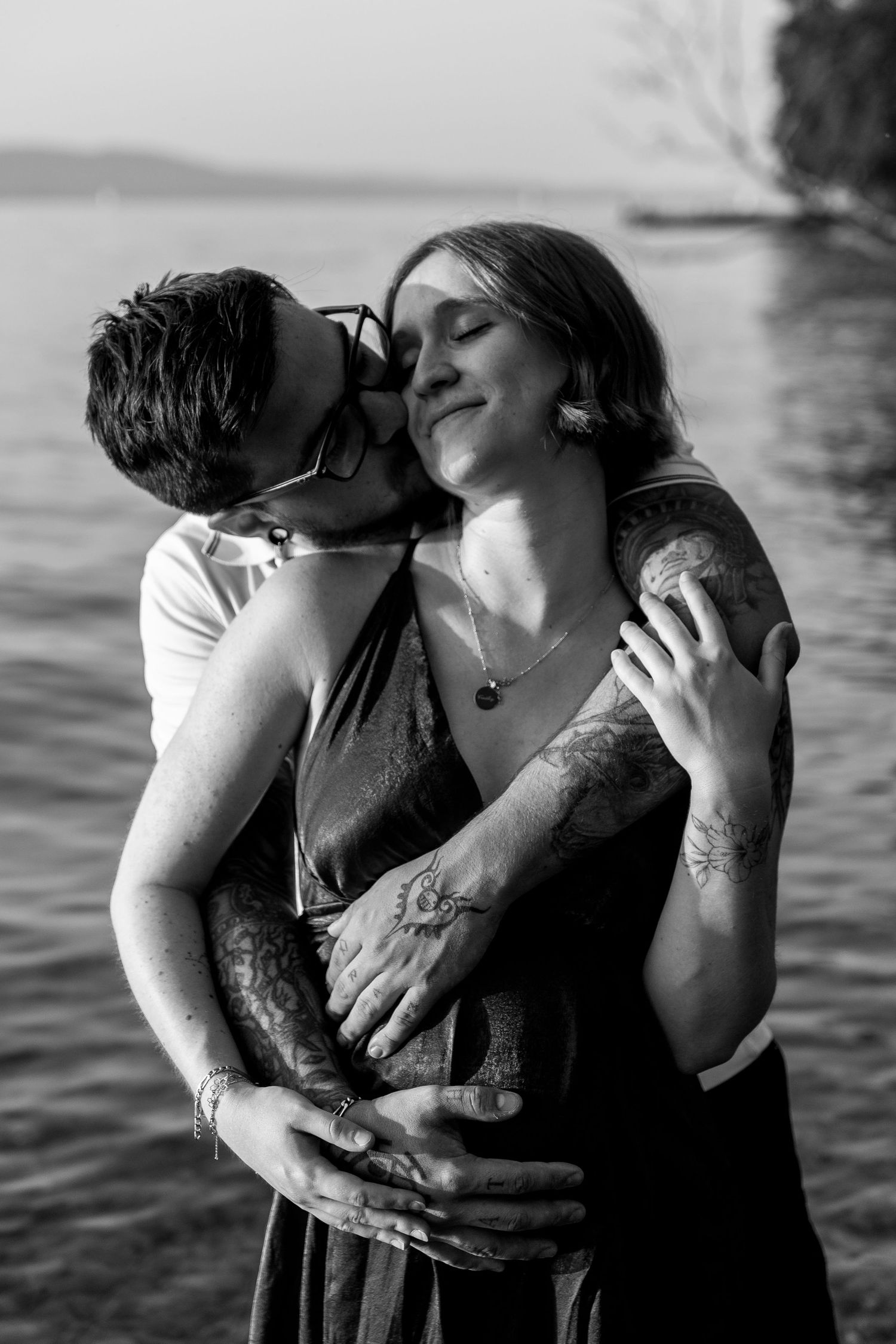Man kisses tattooed pregnant woman's cheek as she smiles blissfully by the water.