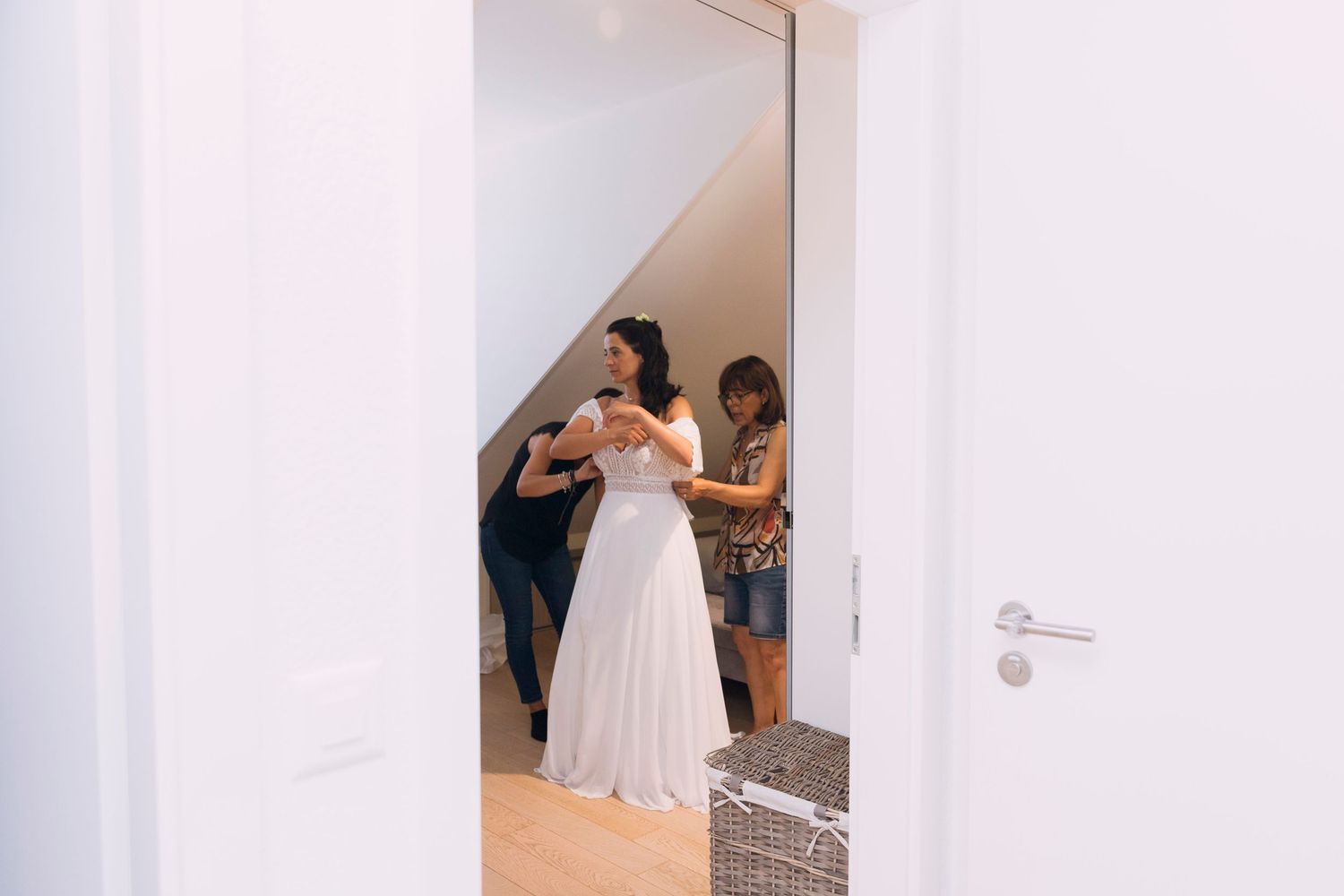 A bride in a white gown being assisted by two women in a bright white dressing room.